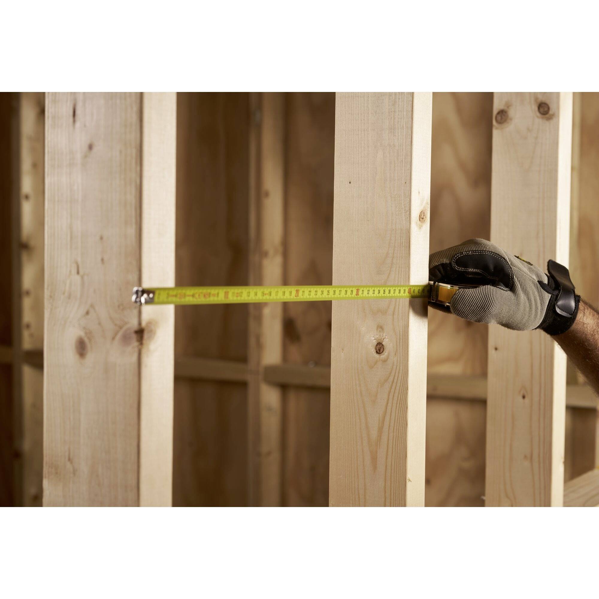 A person is measuring the distance between two vertical wooden beams with a tape measure. They are wearing a glove on their right hand.