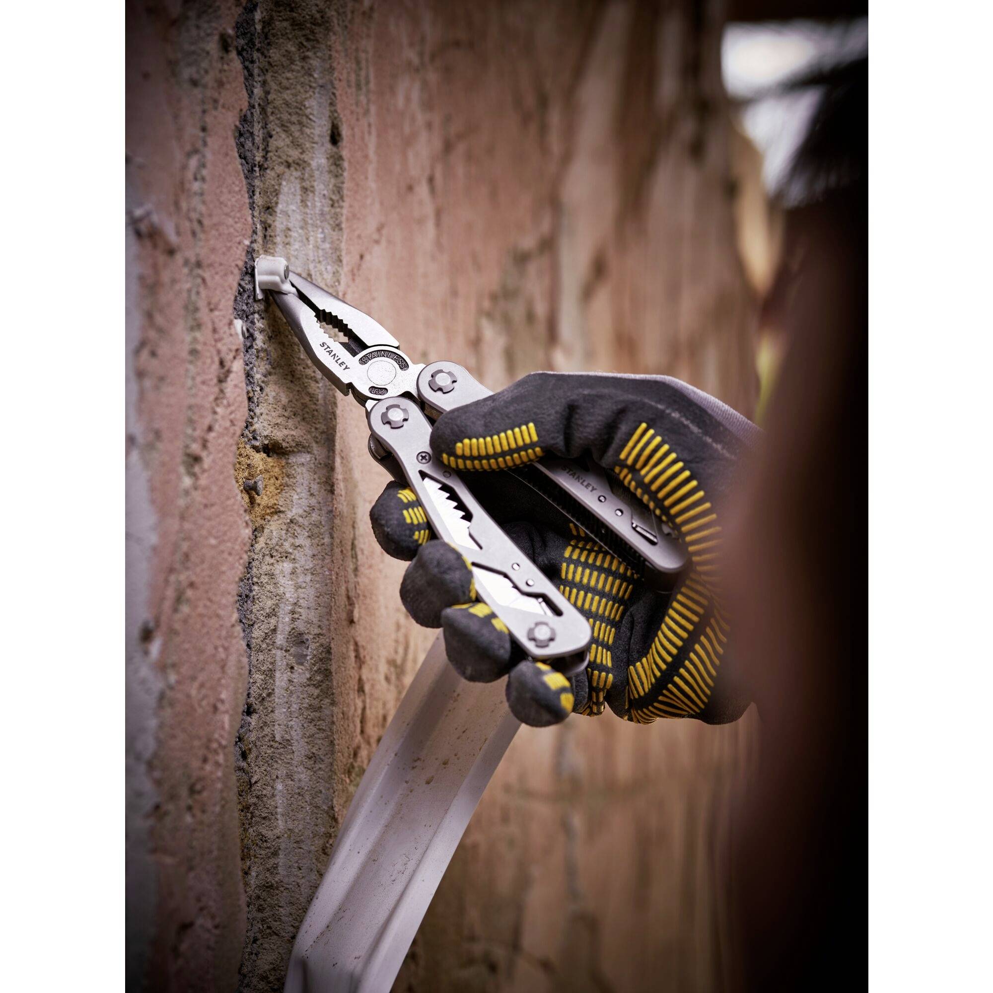 A hand wearing a work glove holds a multi-tool and works on a piece of cable against a brick wall.