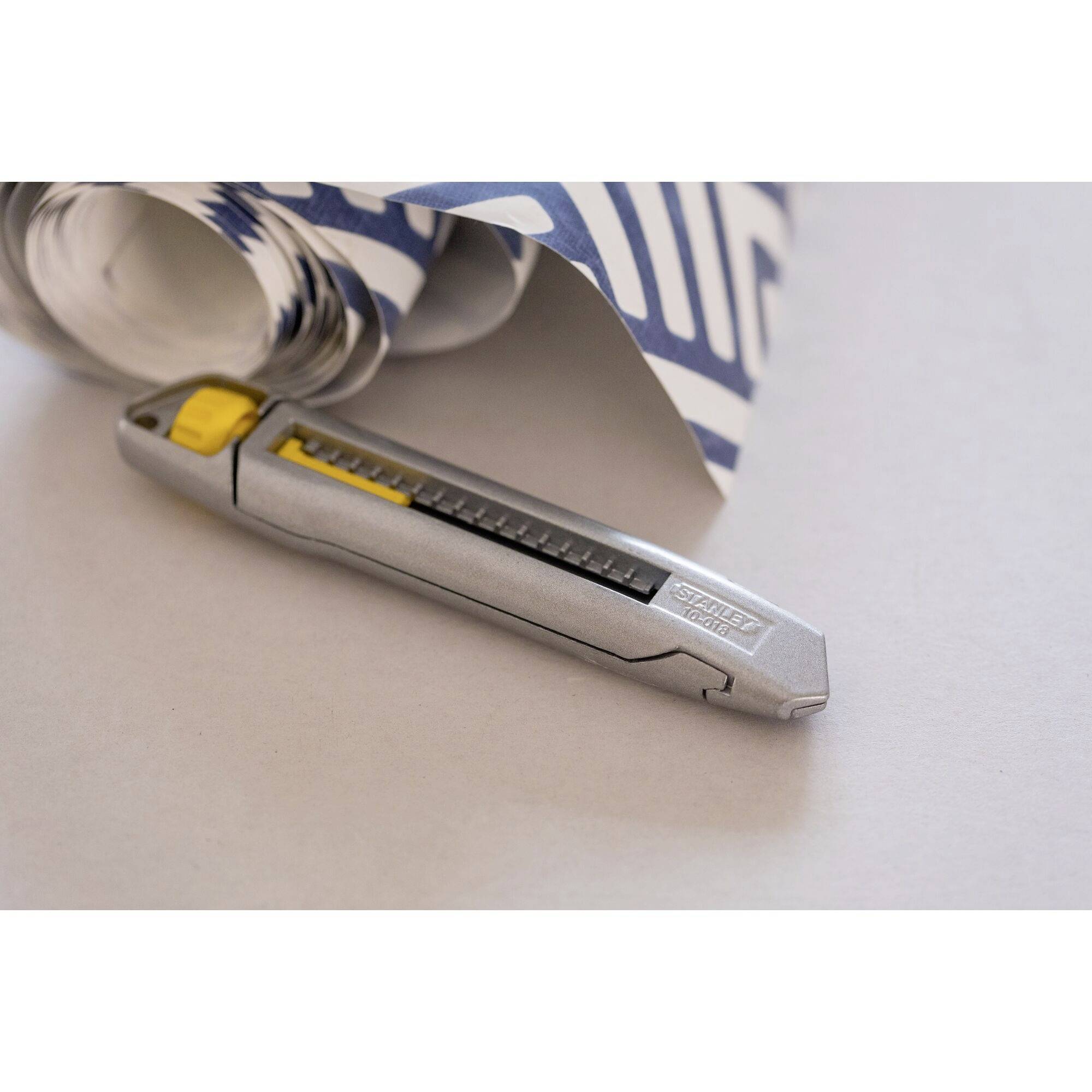 A silver universal cutter lies beside a roll of blue and white patterned wallpaper on a table.