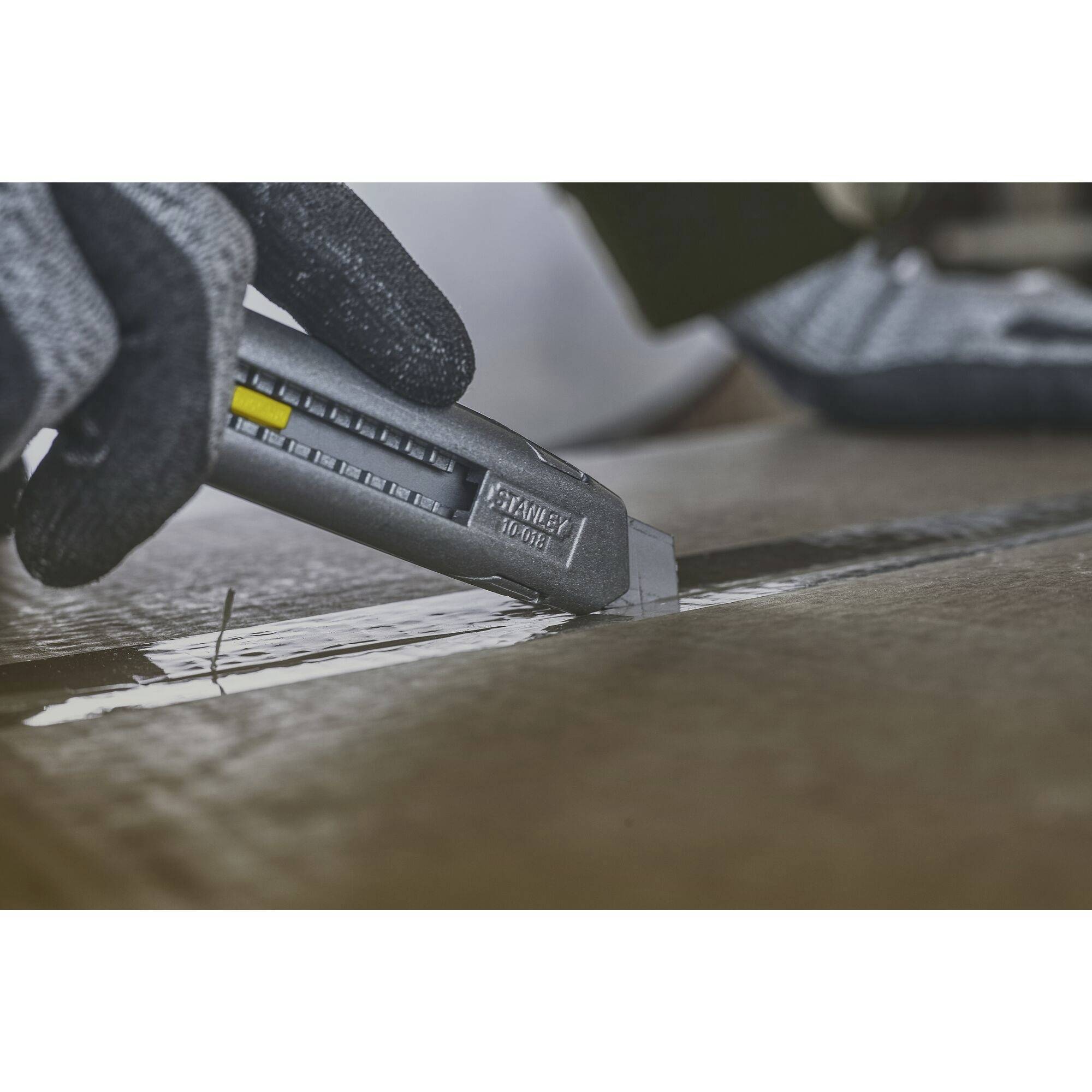 A gloved hand cuts open a cardboard box with a craft knife. The knife has a scale on its side.