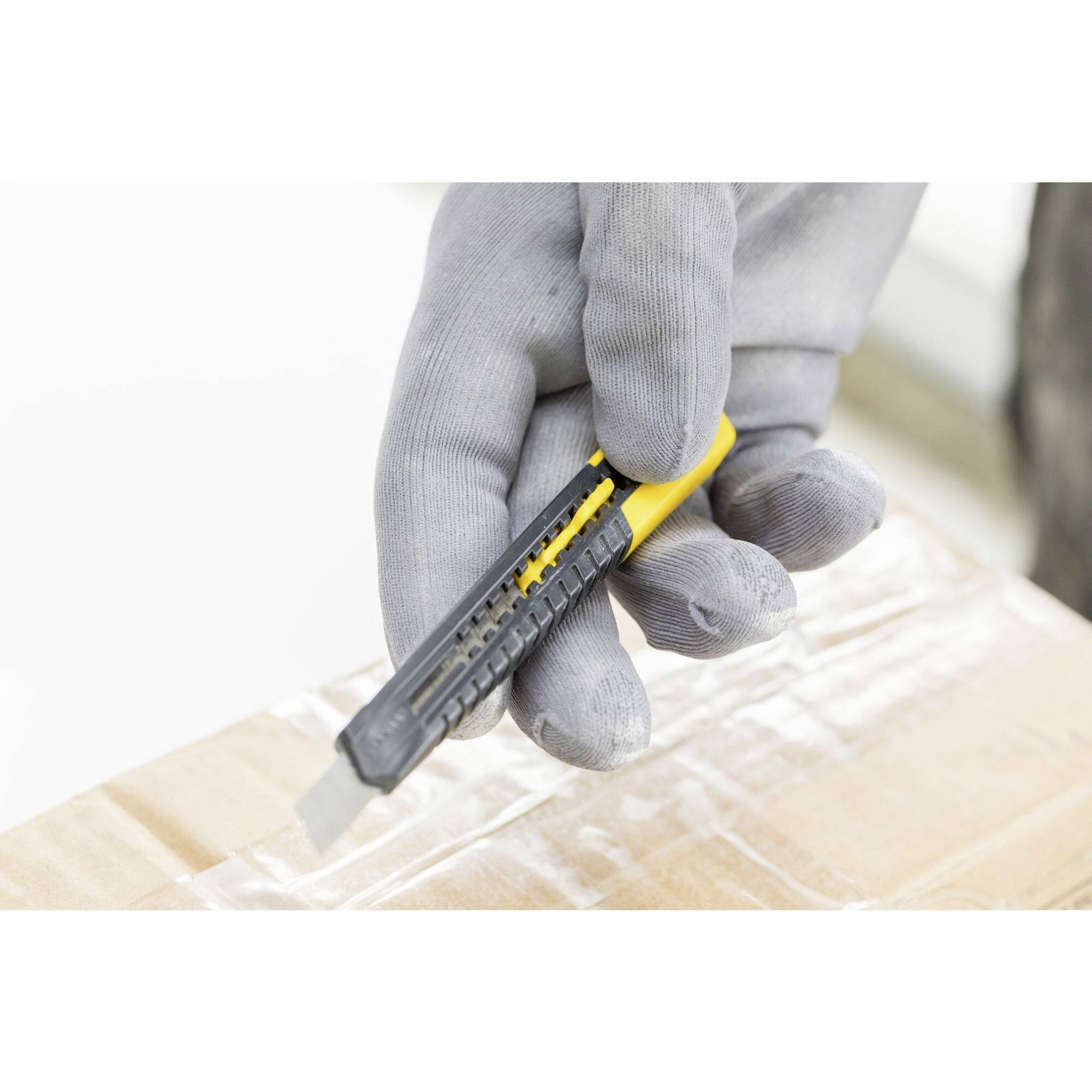 A gloved hand holds a yellow-black utility knife and cuts adhesive tape on a brown cardboard box.