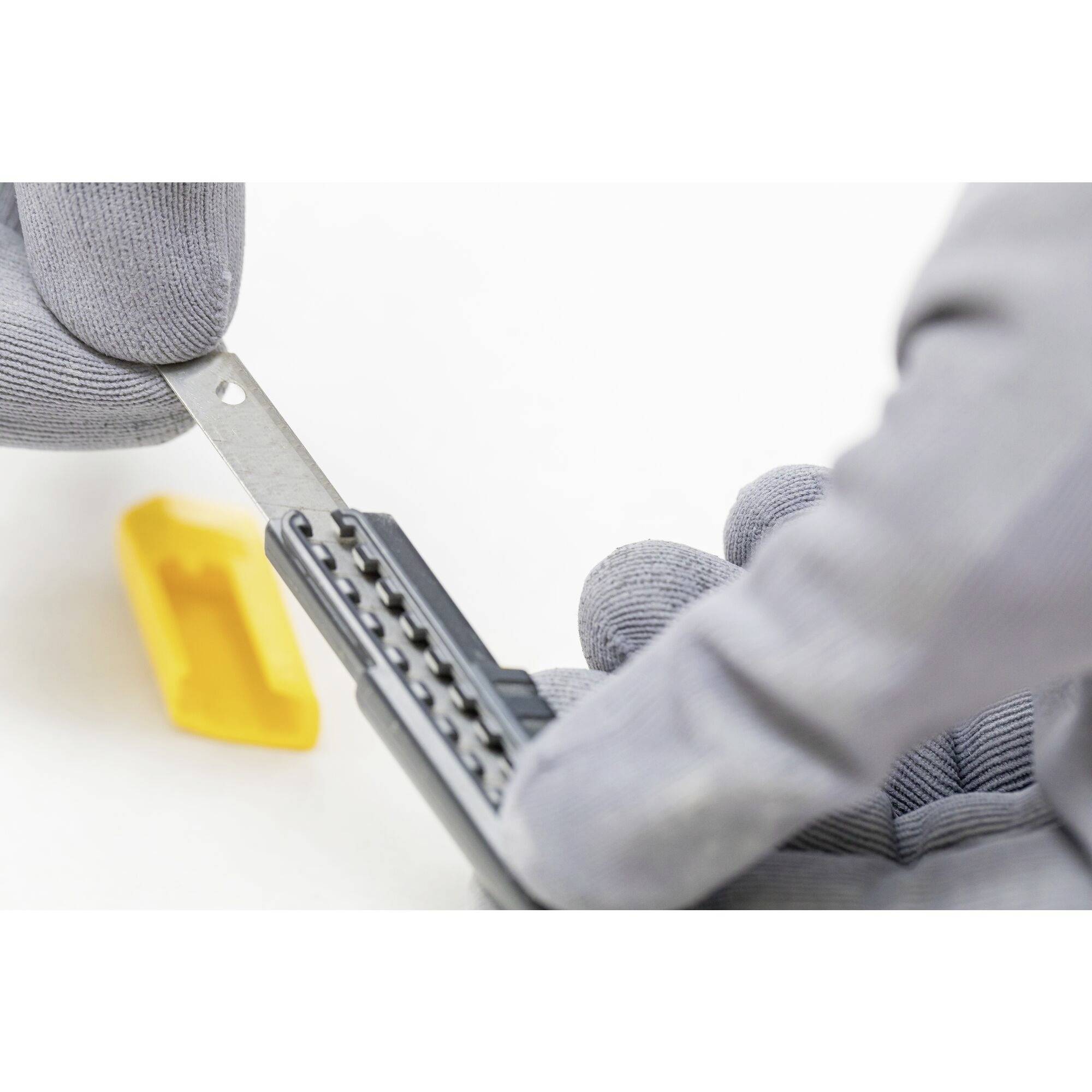 A person wearing grey gloves is holding a metal object with black control points, with a yellow plastic component lying beside it.