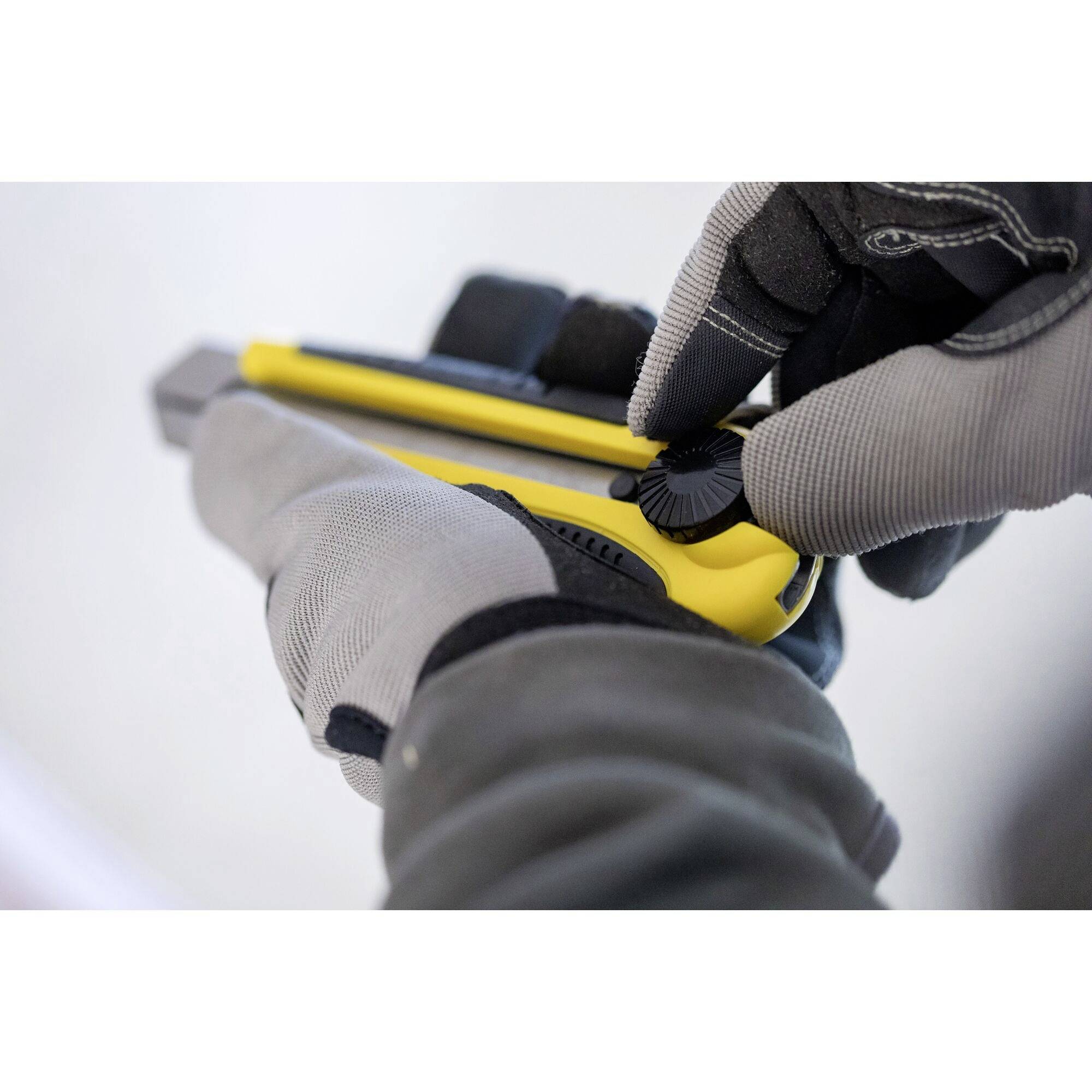 A person is wearing grey gloves and holding a yellow craft knife with a black handle. They are adjusting the blade using the wheel.