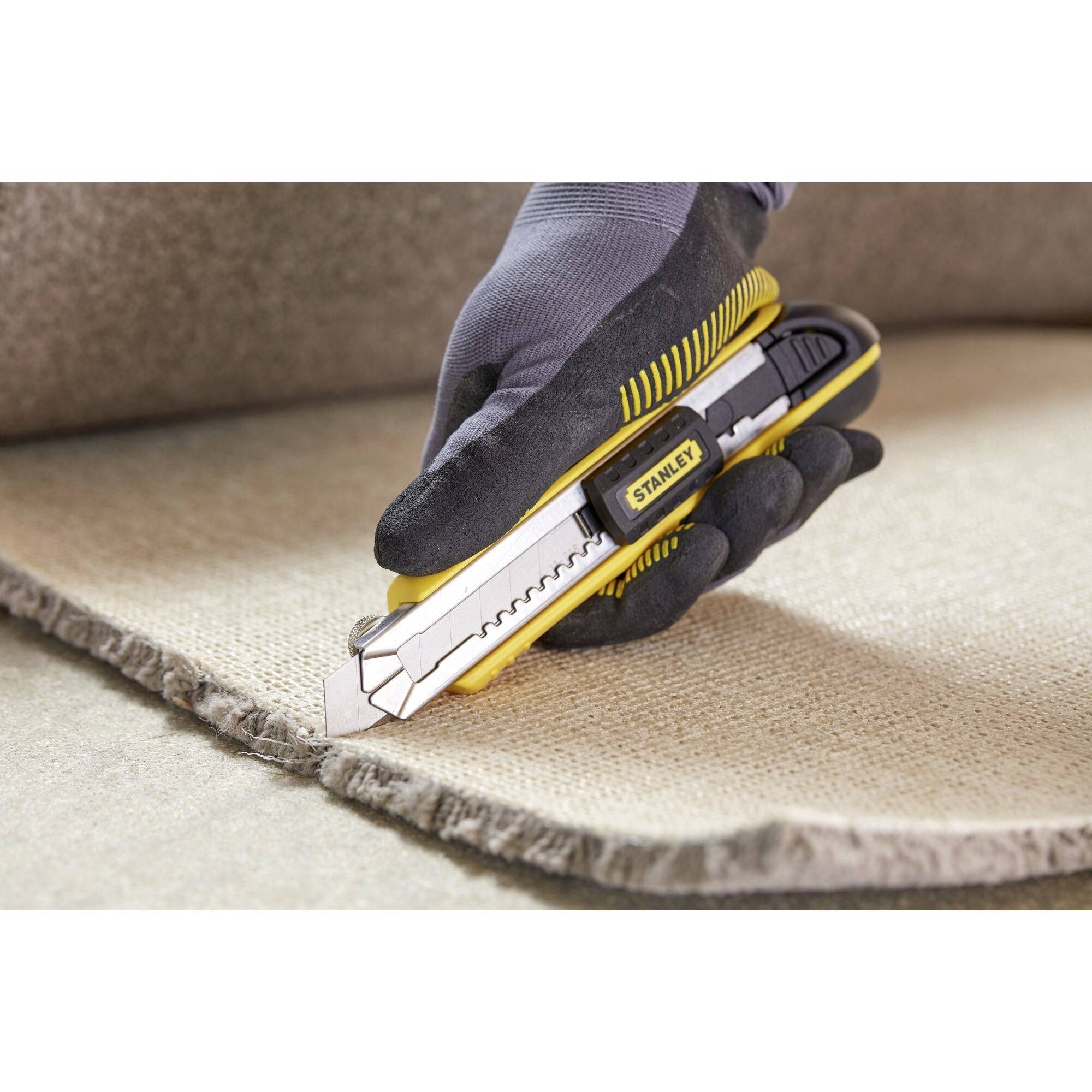 A gloved hand cuts a carpet with a yellow carpet knife along its edge.