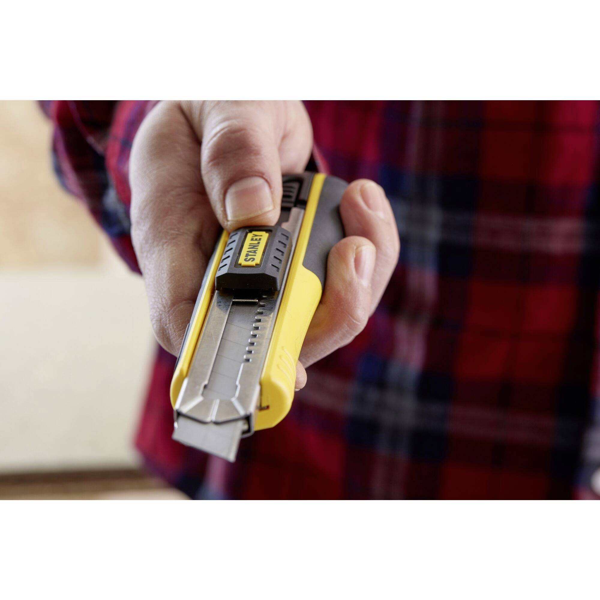 A hand is holding a yellow carpet knife, ready to cut. The person is wearing a checked shirt.