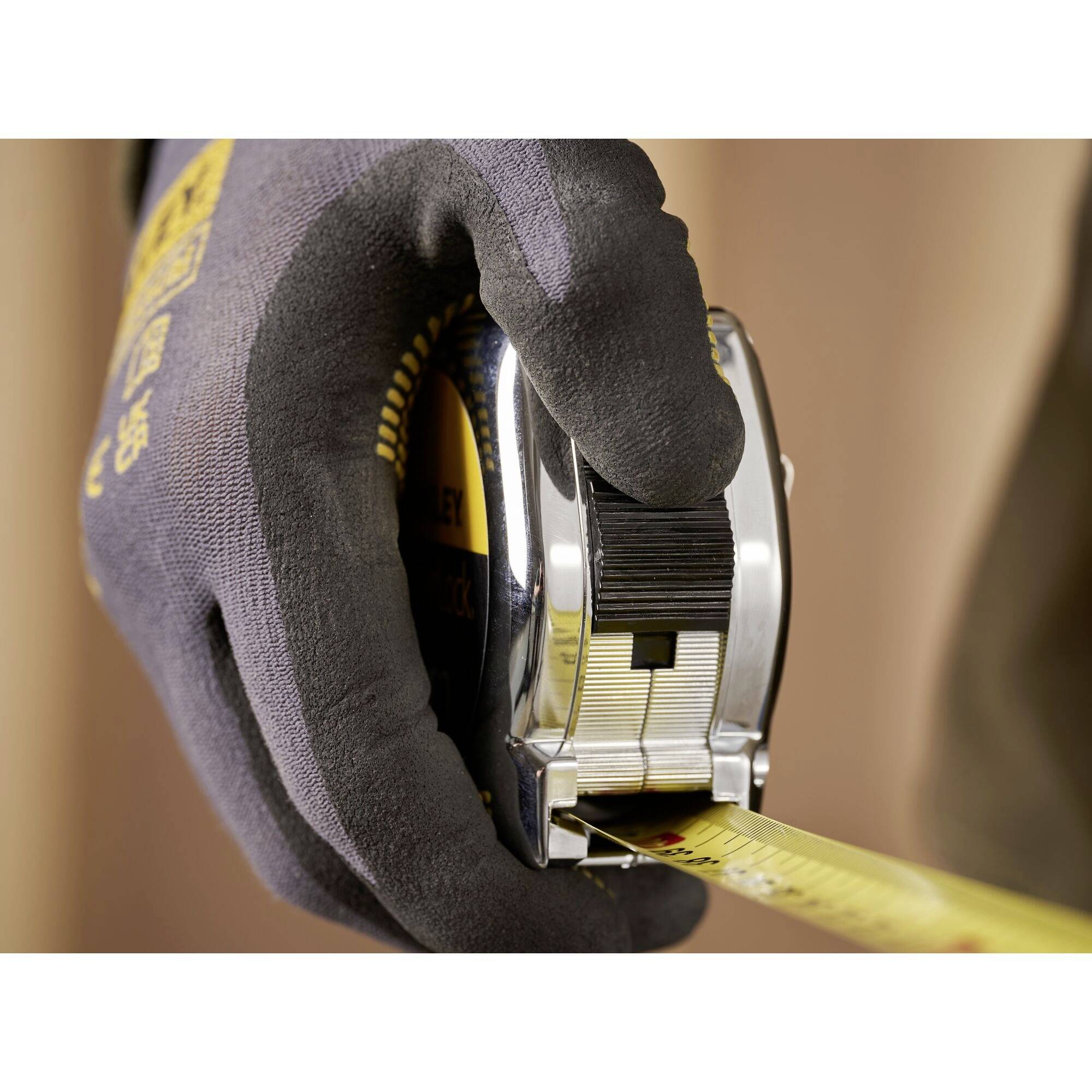 A hand wearing a work glove holds a tape measure that is rolled up. The thumb is pressing the lock to fix the tape in place.