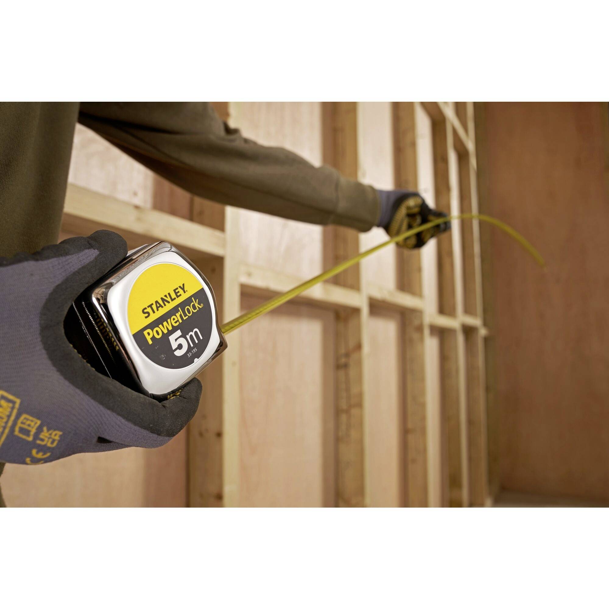 A person is measuring the distance between wooden studs in a construction project using a yellow 5-metre Stanley measuring tape.