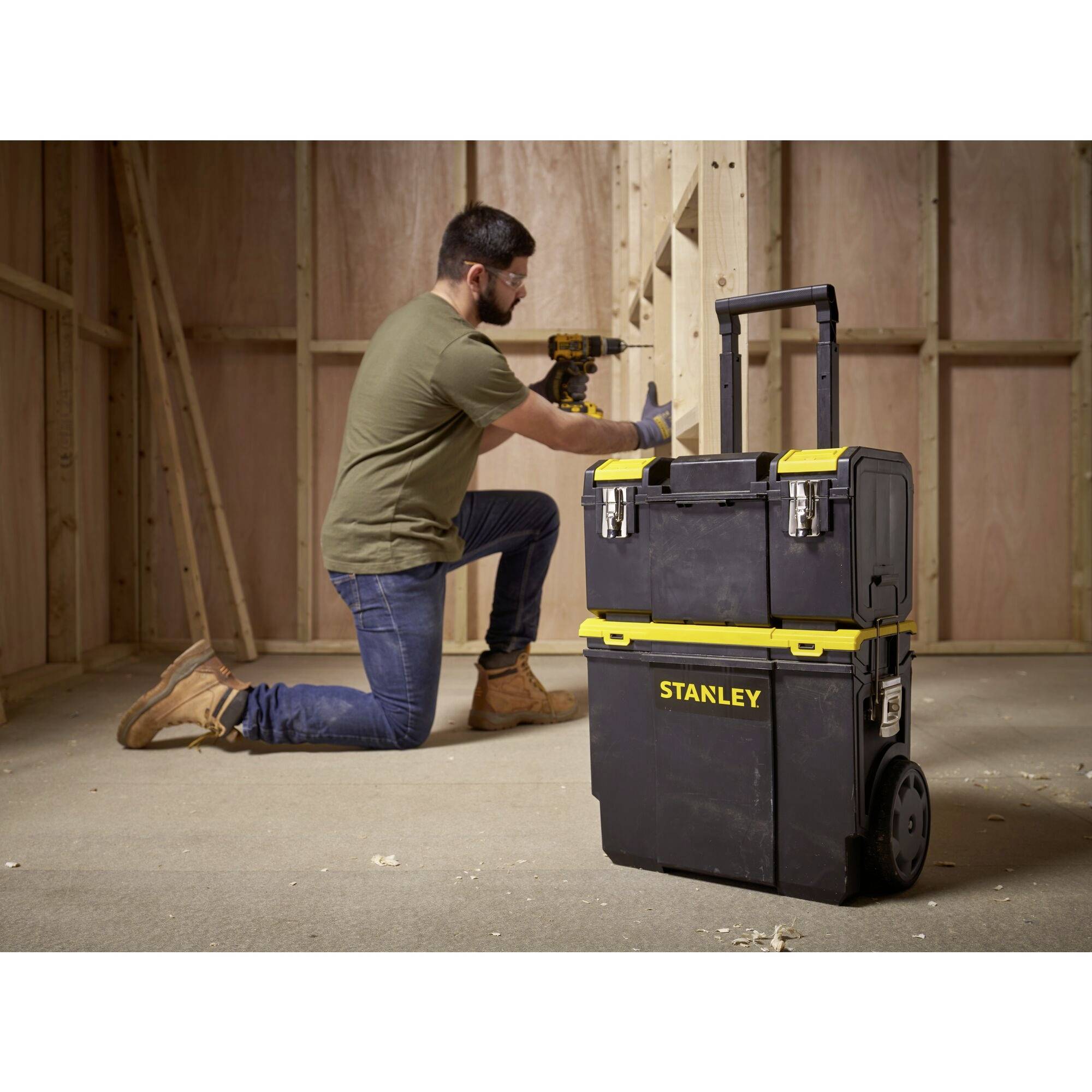 A man in workwear is using a cordless drill in an unfinished room. A large toolbox on wheels stands in the foreground.