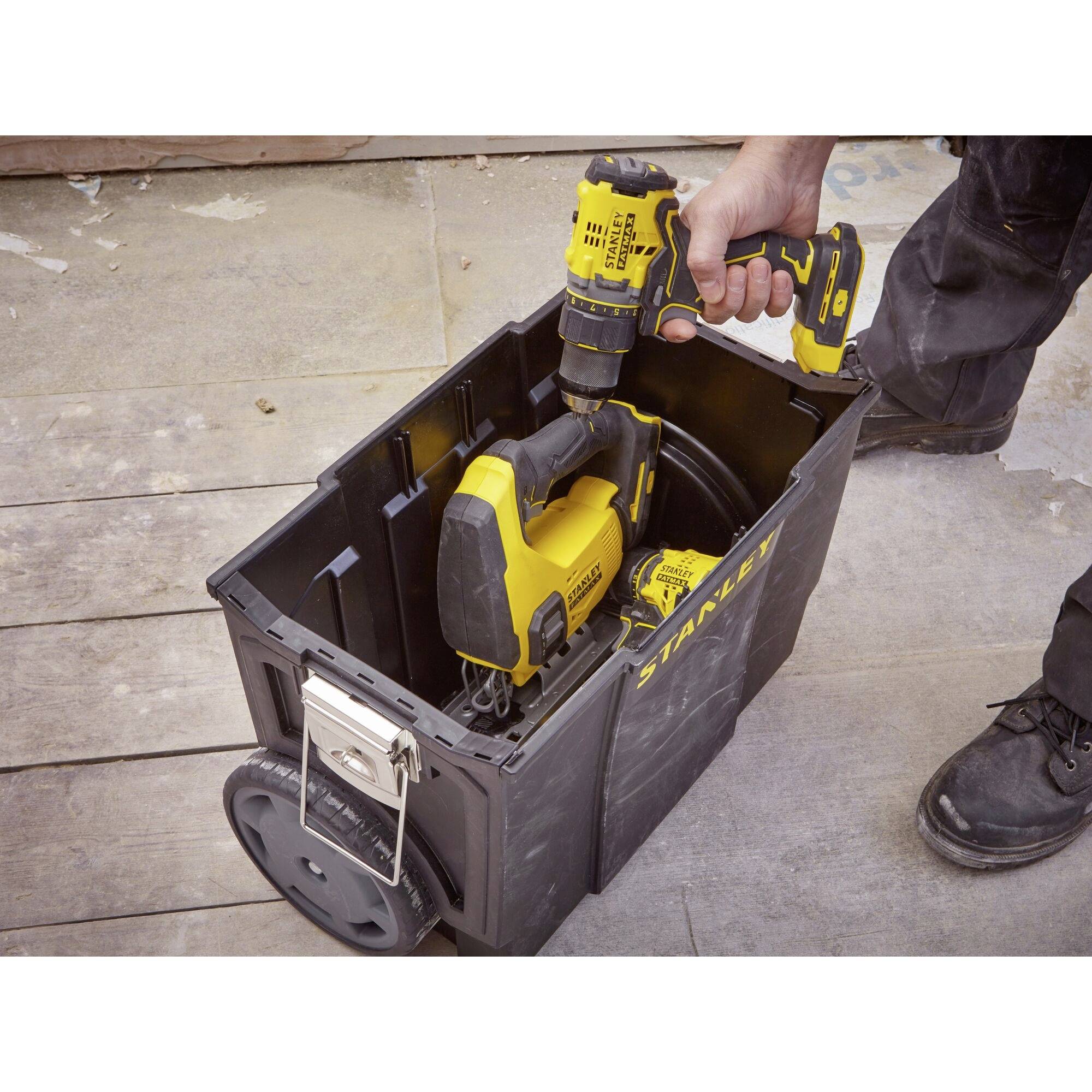 A hand is reaching for a yellow drill from an open black toolbox situated on a wooden floor.