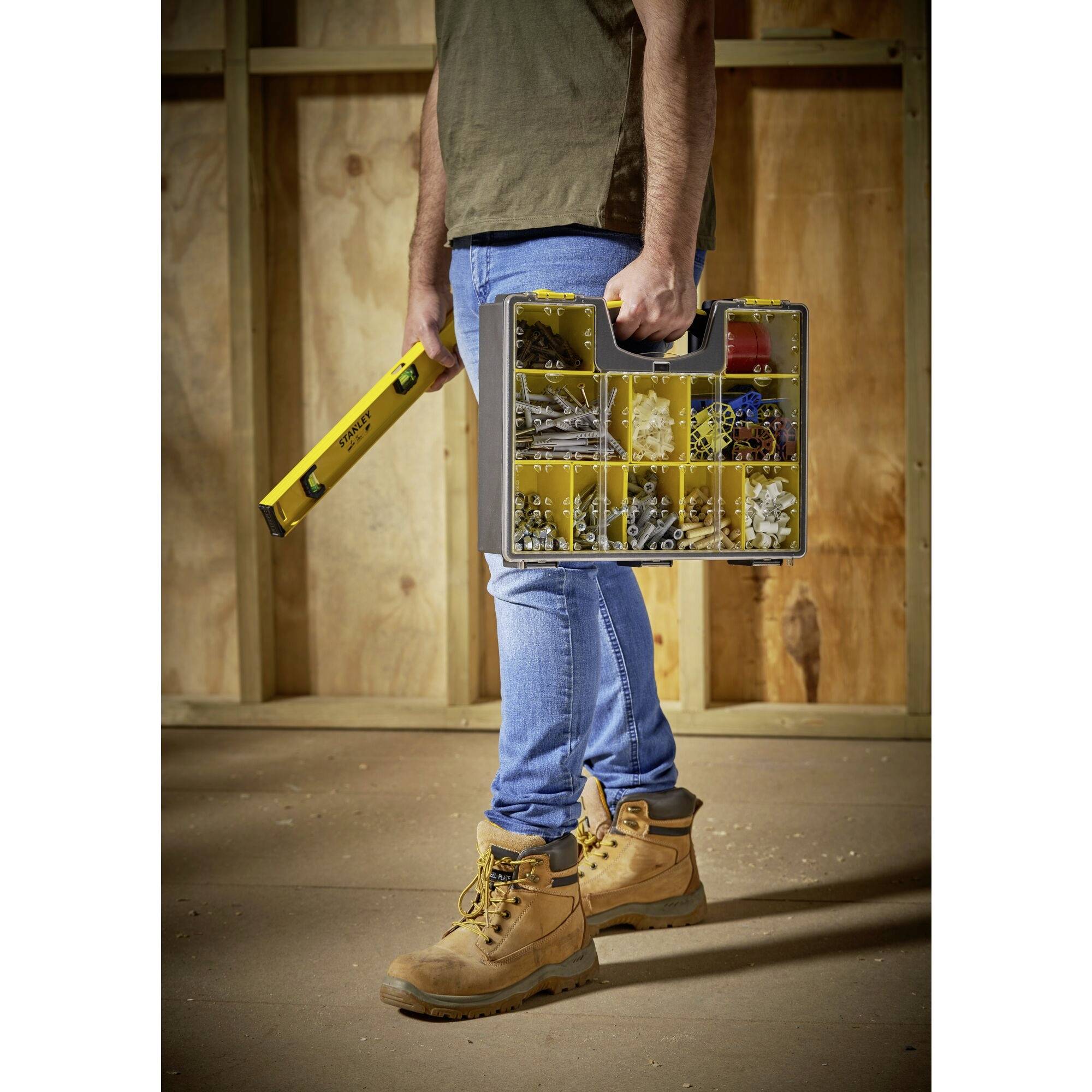 A person is carrying a toolbox with various screws and nails, along with a spirit level. They are standing in an unfinished room.