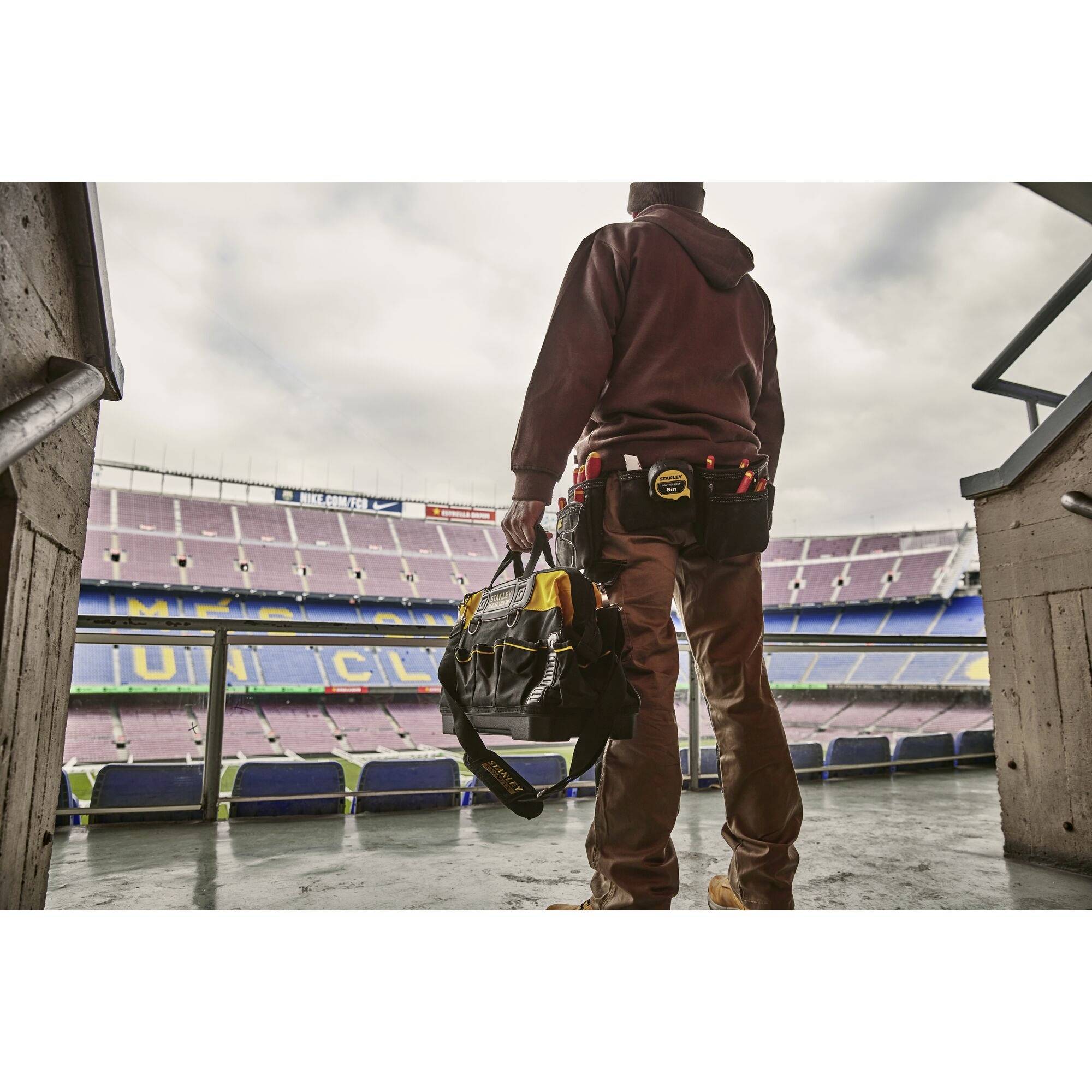 A person in work attire stands at the entrance of an empty football stadium, holding tool bags.