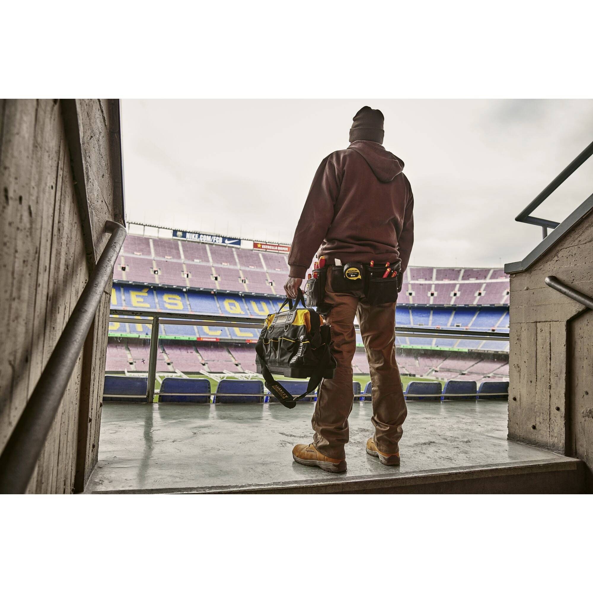 A person in workwear stands with a bag in the football stadium and looks out over the pitch.