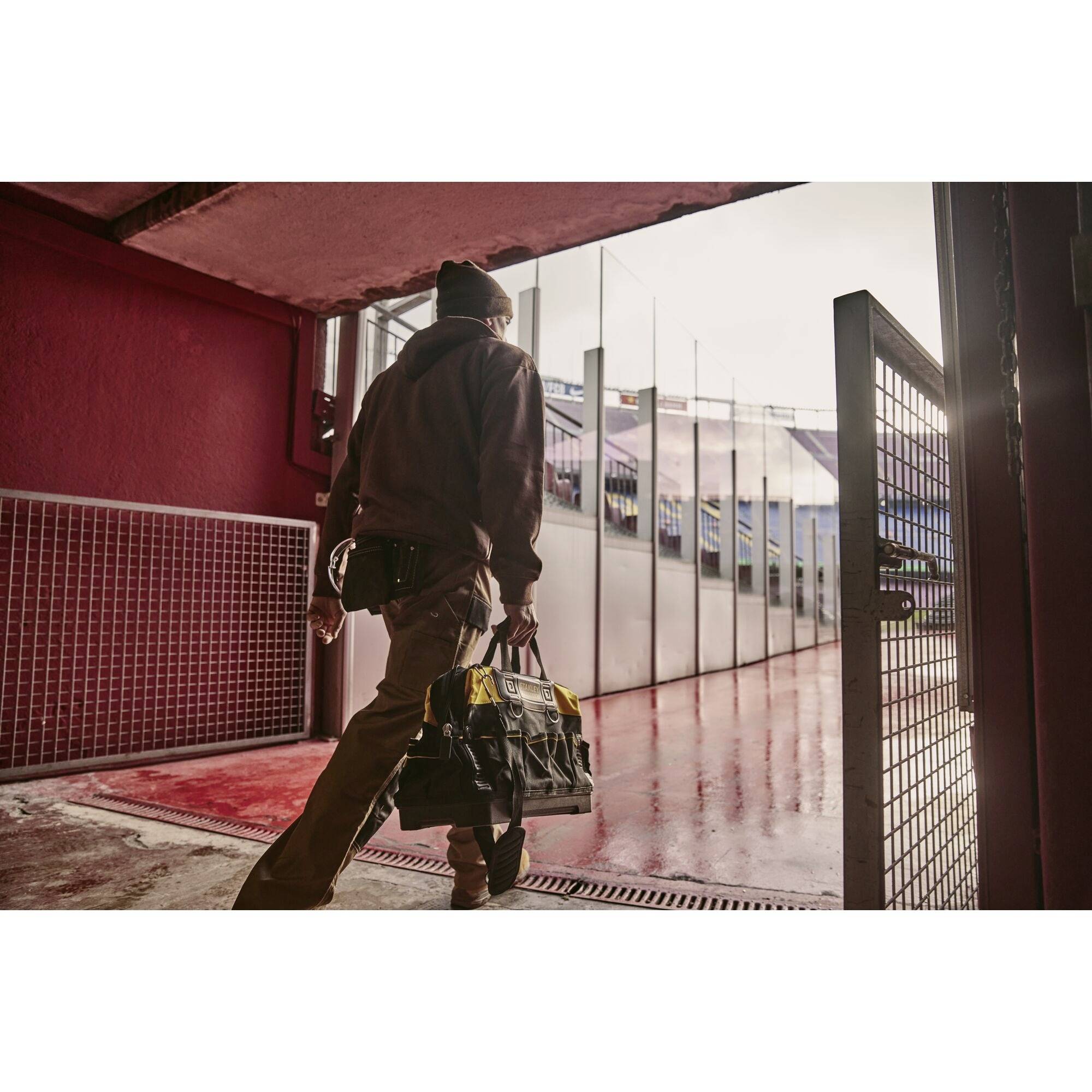 A person in workwear is carrying a large bag through a gate in a stadium. The ground is red, and it is cloudy.