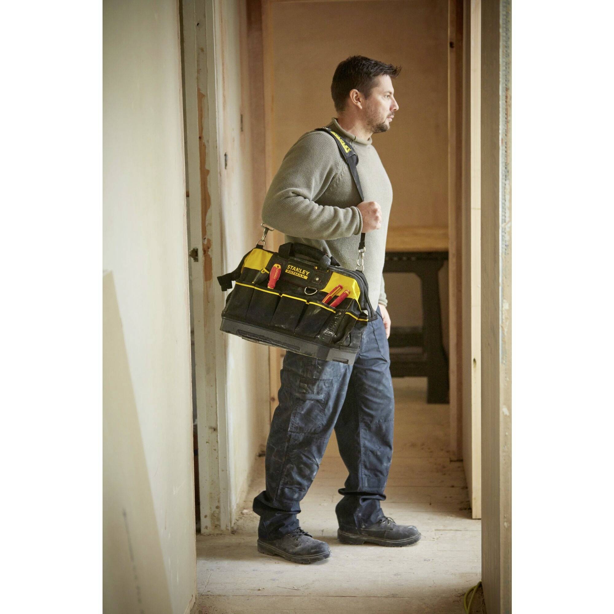 A man stands in a room, carrying a large tool bag full of tools over his shoulder, gazing into the distance.