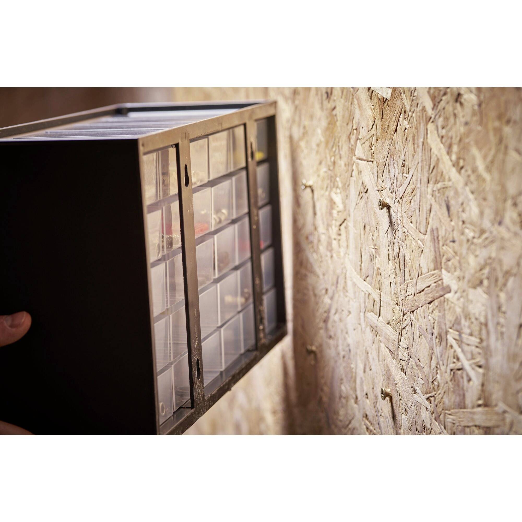 A black storage box with transparent drawers is mounted on an OSB board. A hand is holding the box.