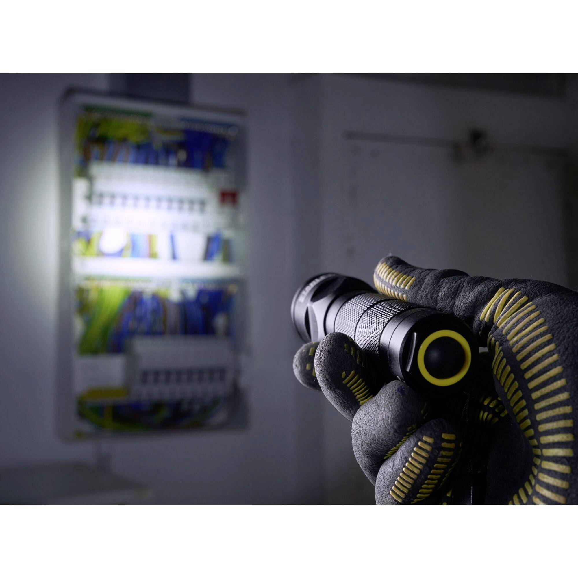 A hand in a work glove holds a torch, shining it onto an open electrical box to illuminate the cables and components.
