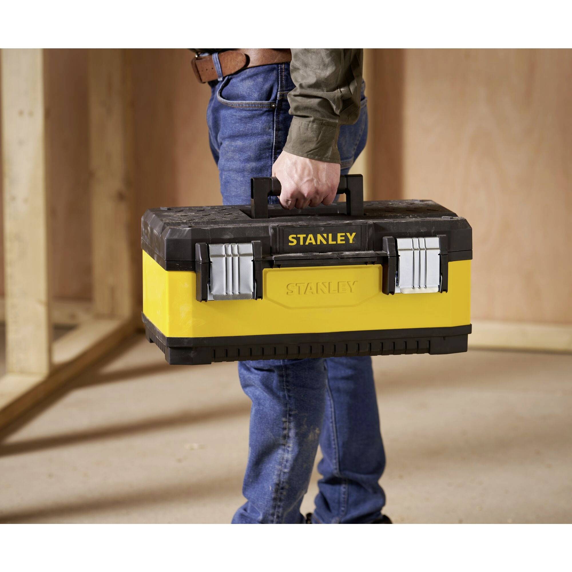 A person is carrying a yellow toolbox in their hand while standing in an unfinished room or workshop. The box is marked with the 'Stanley' label.