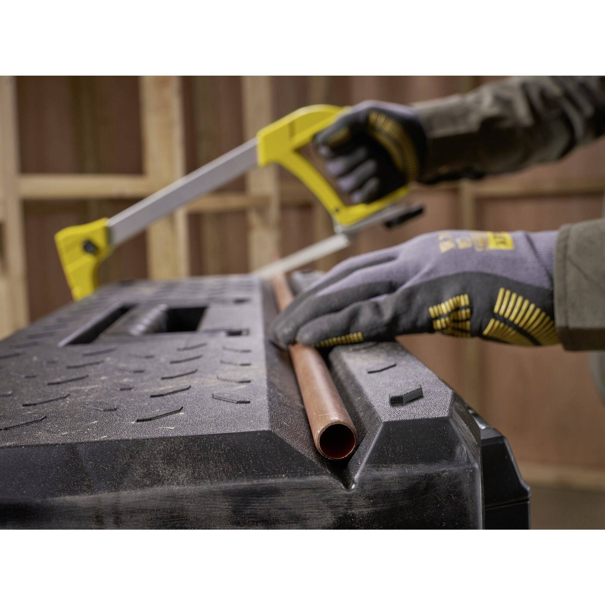 A person wearing gloves is cutting a copper pipe with a hacksaw on a workbench.