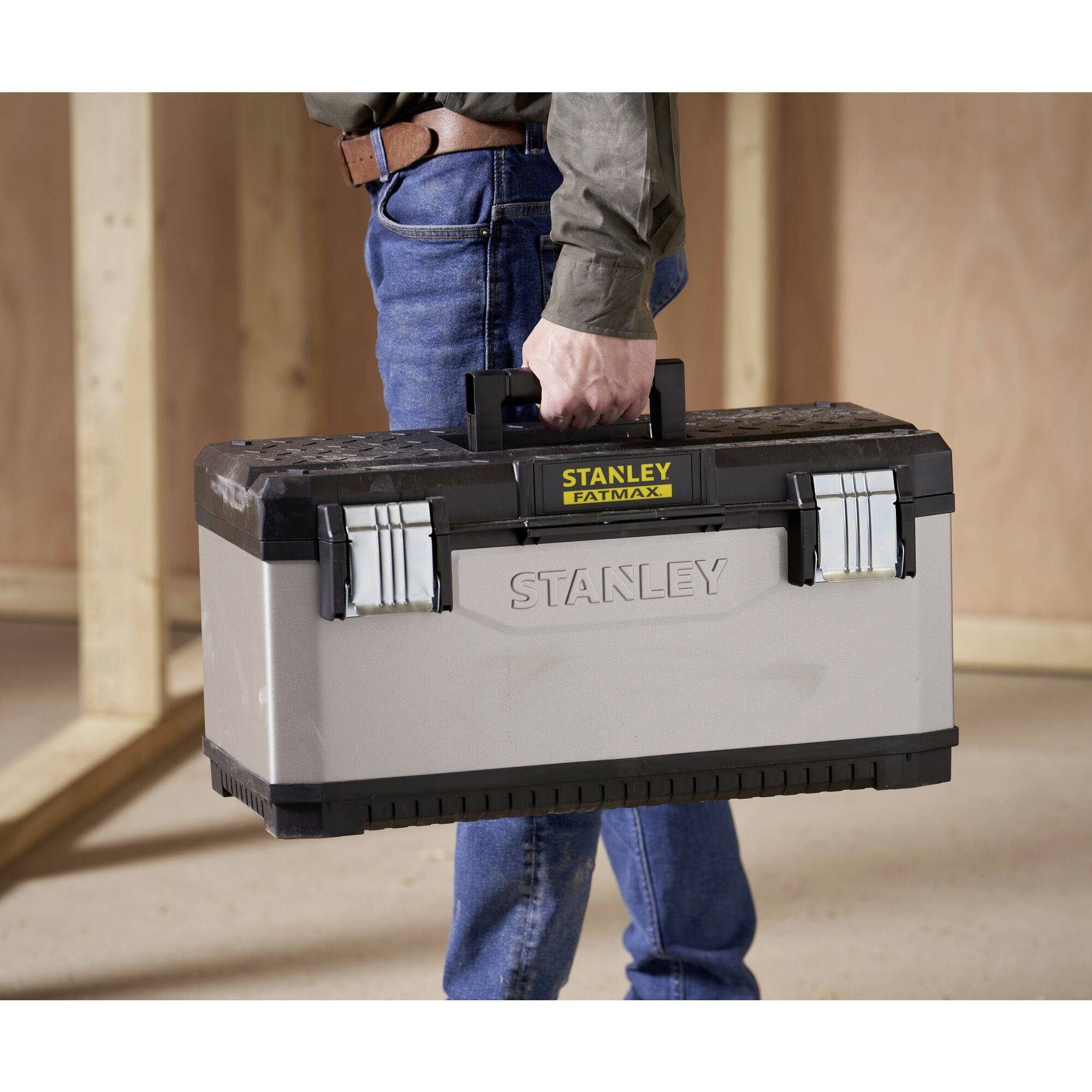 A person is carrying a large, grey toolbox with 'STANLEY FATMAX' written on it in a construction site environment.