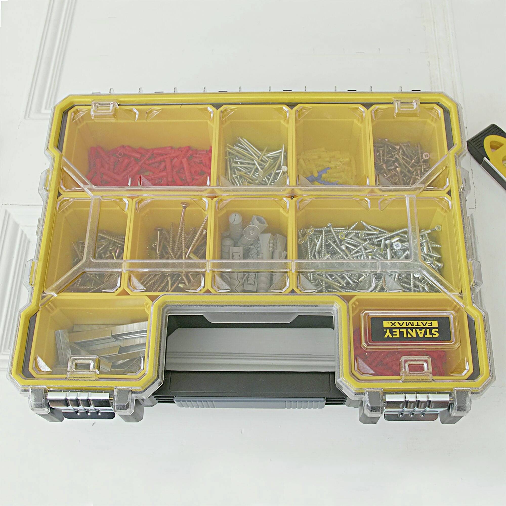 Yellow toolbox with a transparent lid, filled with various screws and wall plugs in separate compartments; a tape measure placed beside it on the right.