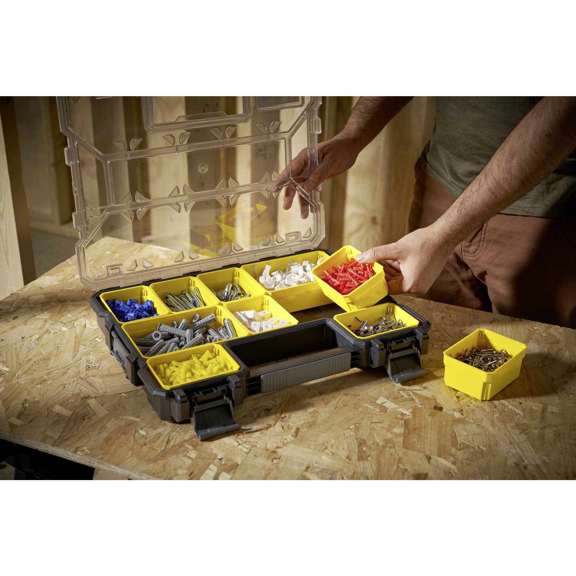 A person is sorting various screws and wall plugs in a transparent box with yellow compartments on a workbench.
