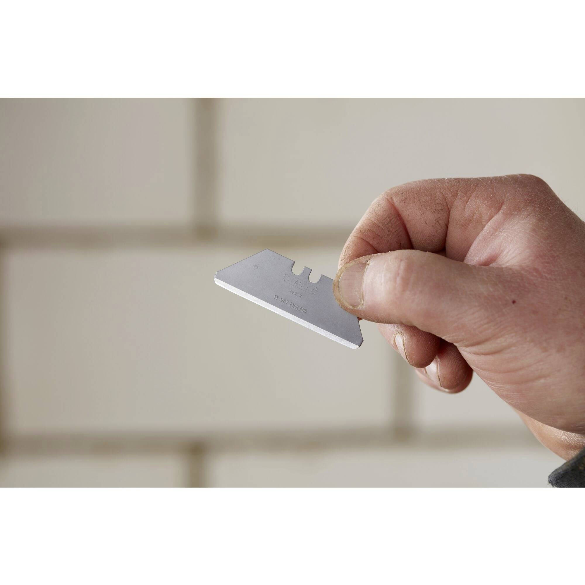A hand holds a razor blade in front of a blurred, brick wall.