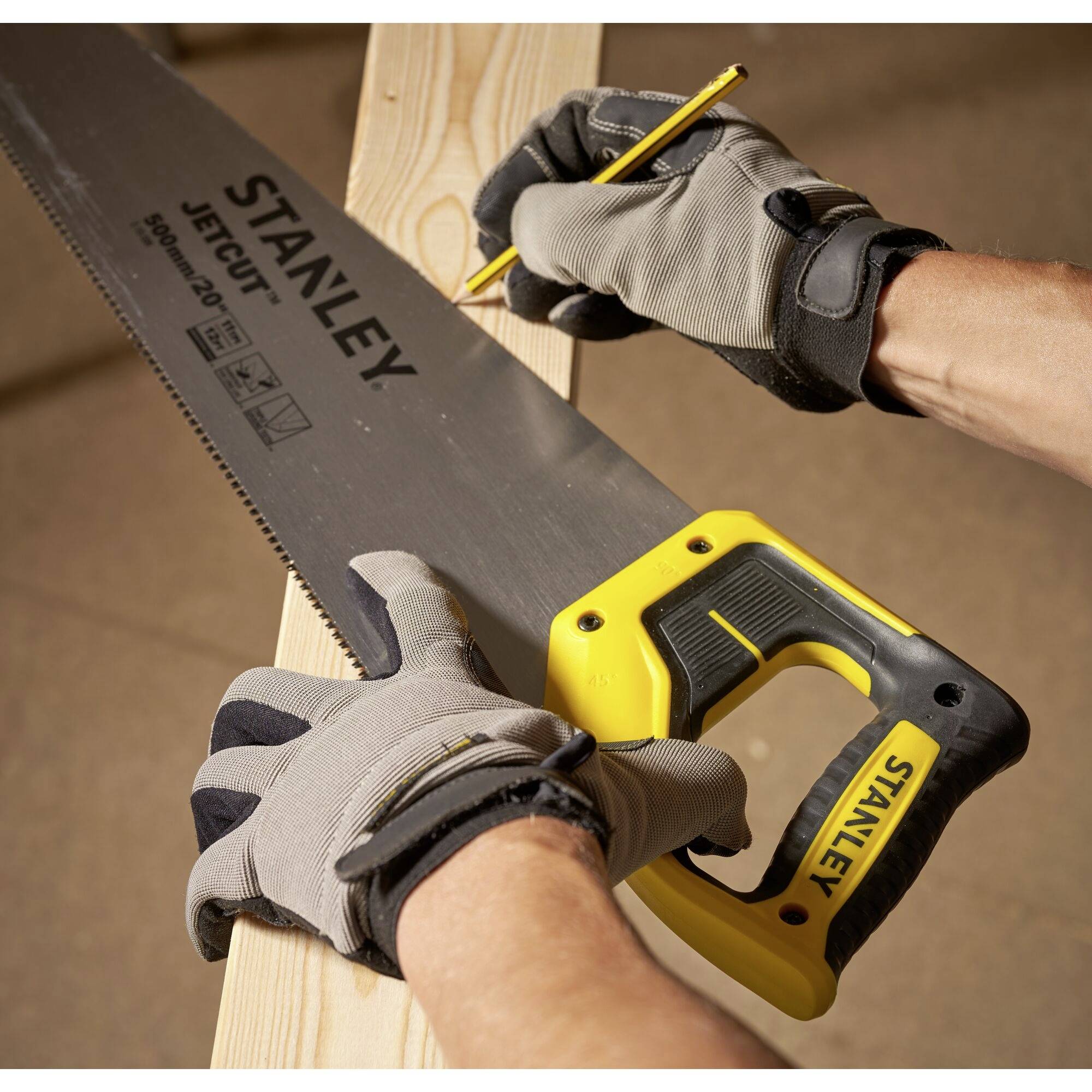 A person wearing work gloves is sawing a wooden board with a Stanley saw while marking it with a pencil.