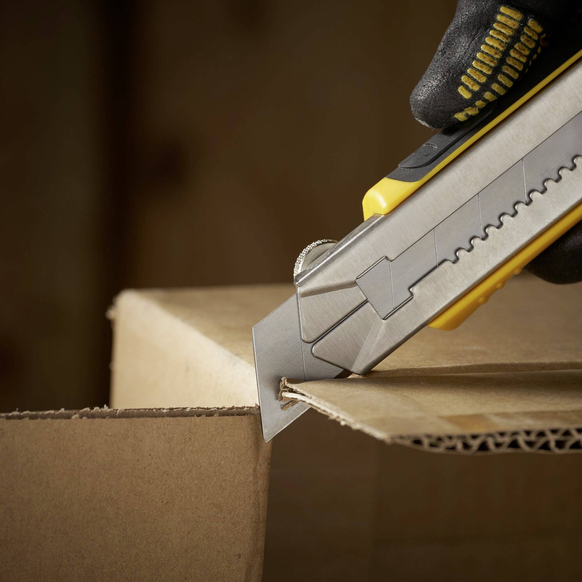 A hand wearing a glove cuts open a cardboard box with a utility knife.