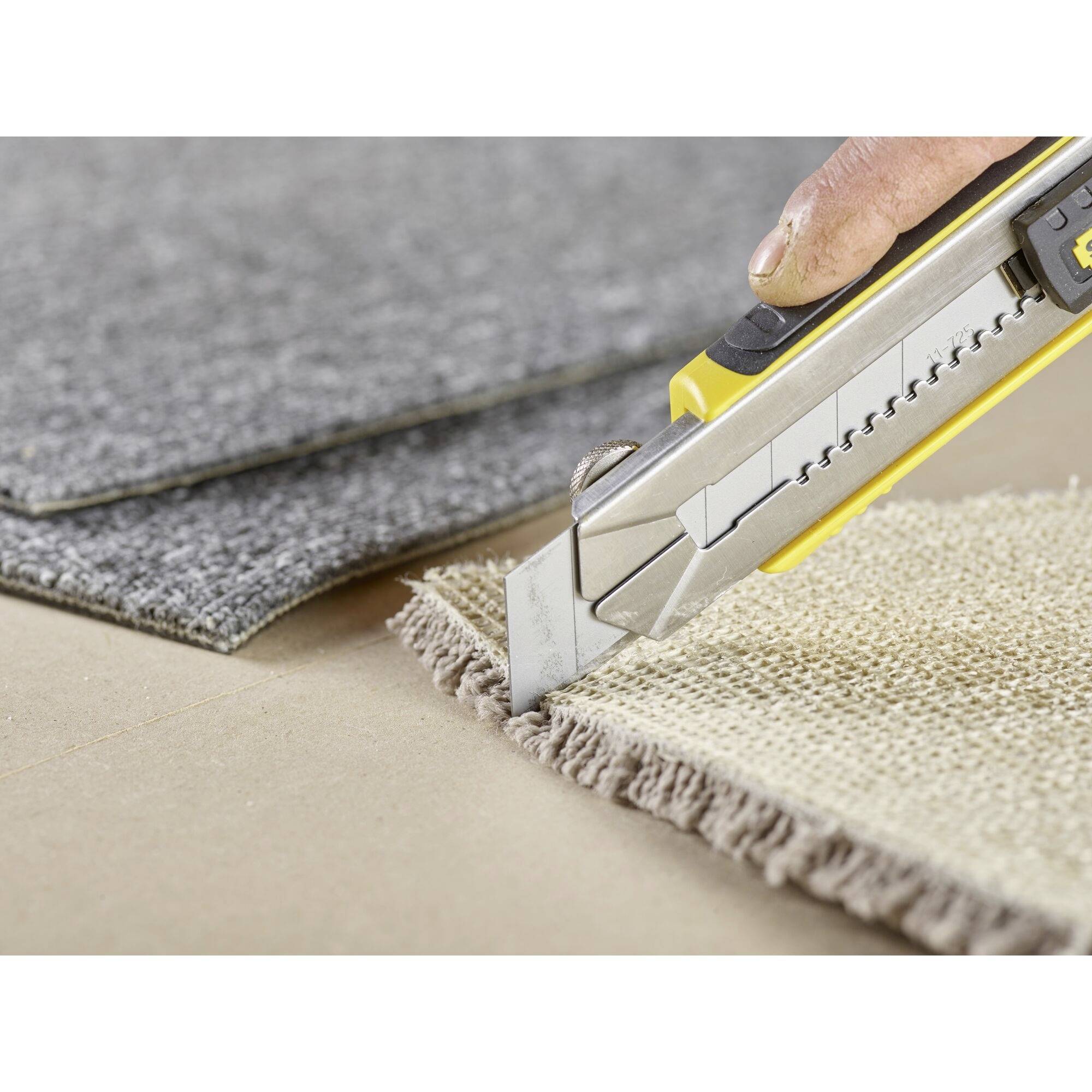 A hand is cutting a carpet with a utility knife on cardboard, with a grey carpet lying beside it.