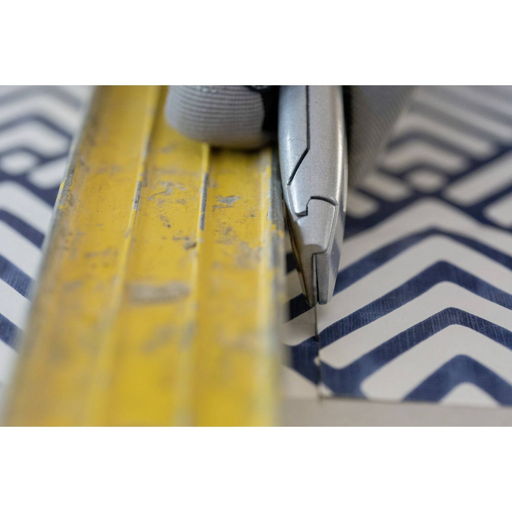 A craft knife cuts along a yellow ruler over a dark blue and white patterned material.