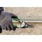 A hand holding a green metal scissors cuts a piece of wire mesh fencing. The background is a beige stone wall.