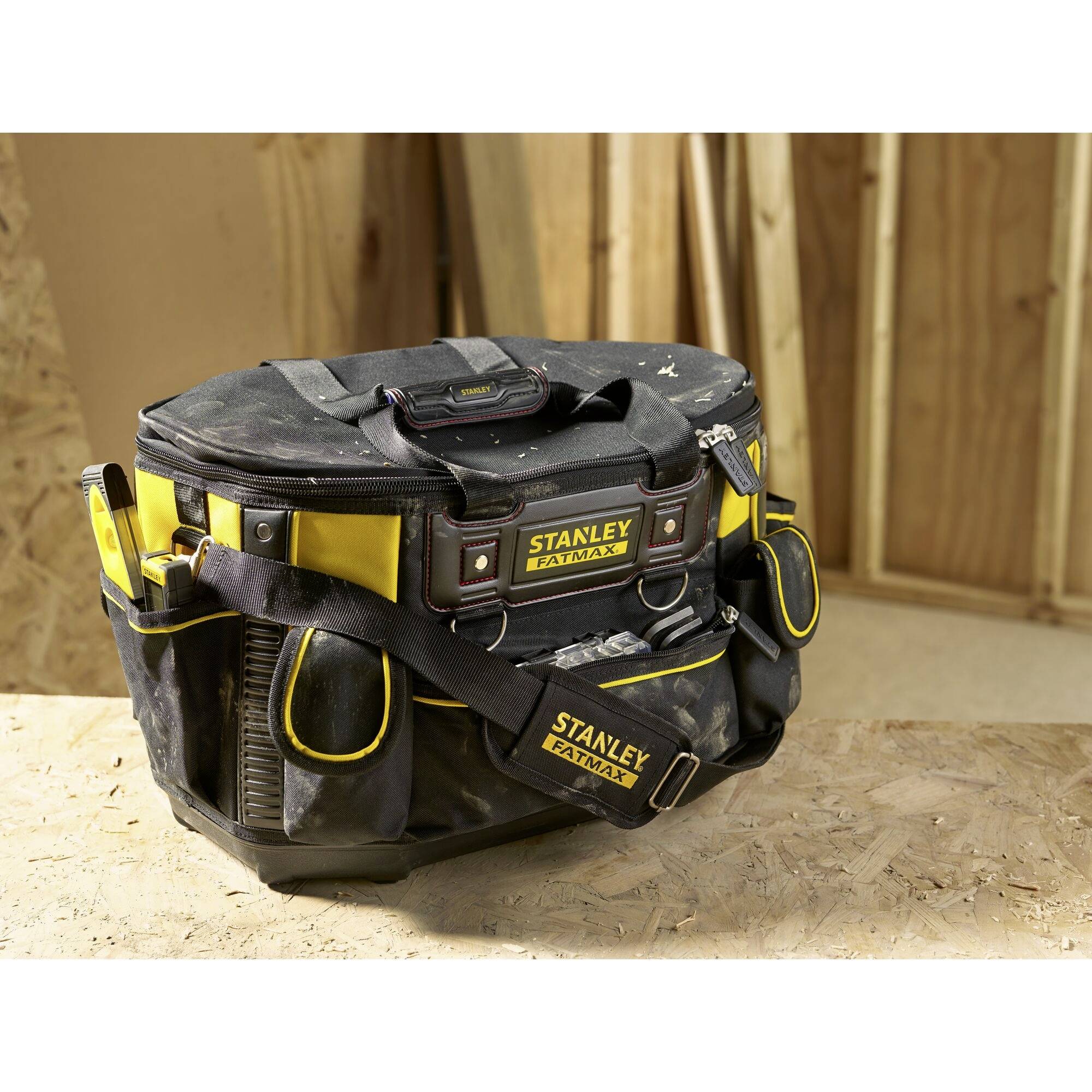 A black tool bag with yellow accents and multiple compartments sits on a wooden table; the background shows wooden walls.