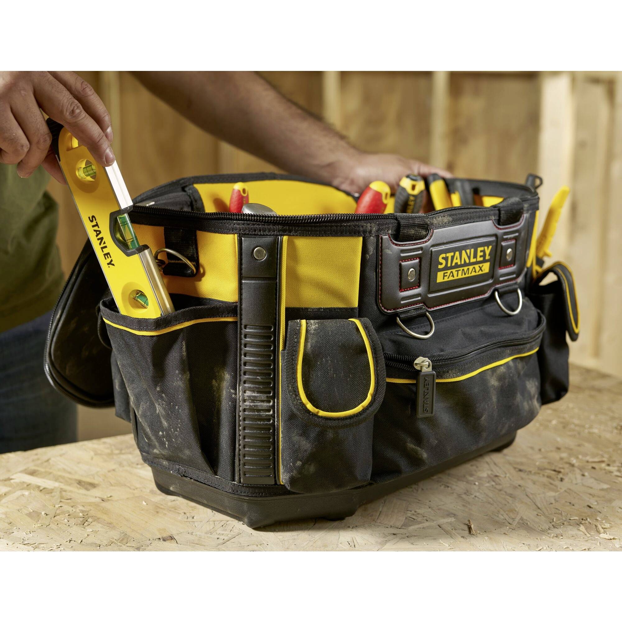 A person is placing a yellow spirit level into a black tool bag on a wooden surface. The bag is filled with tools.