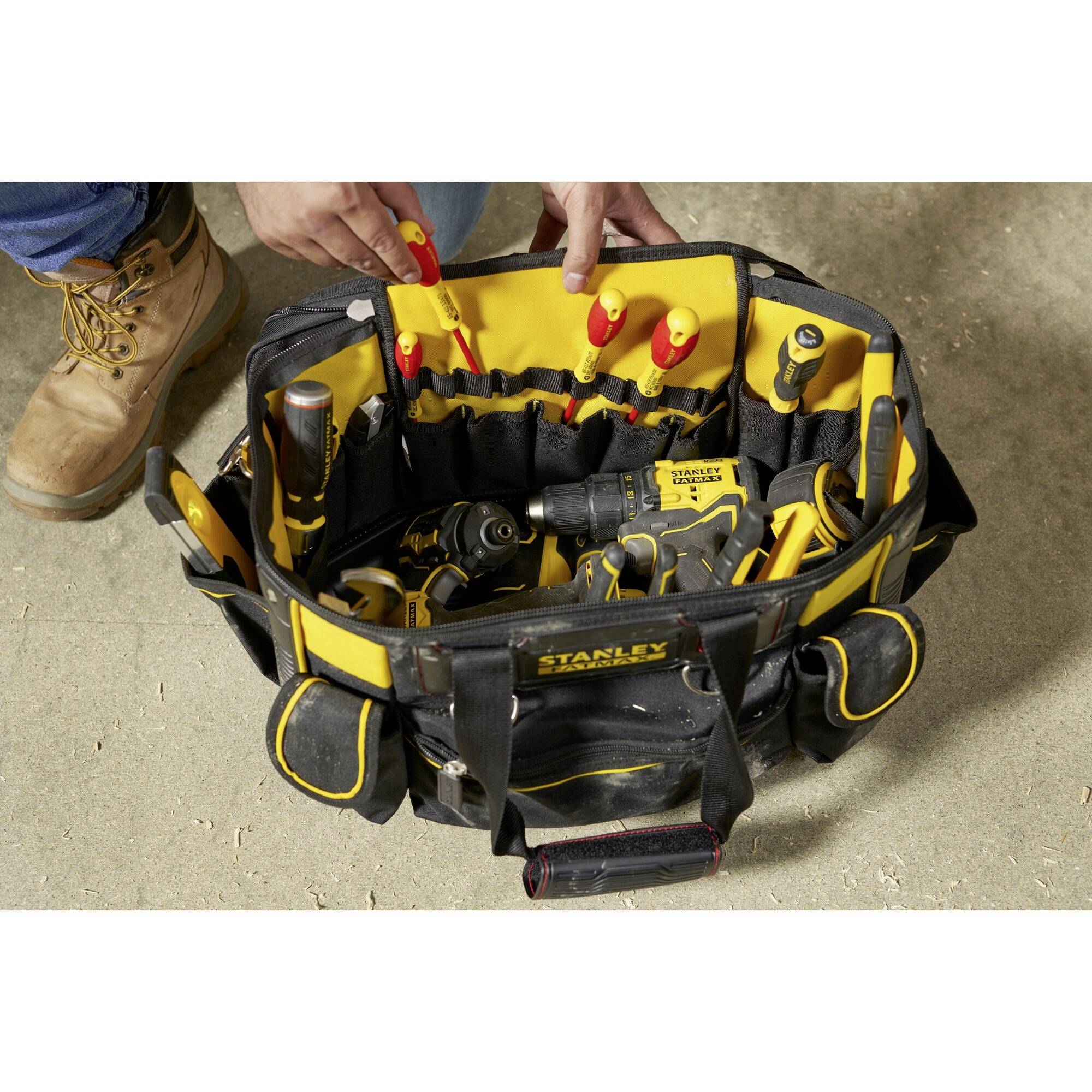 An open toolbag on the floor, filled with various hand tools and power tools. Hands reaching into it.
