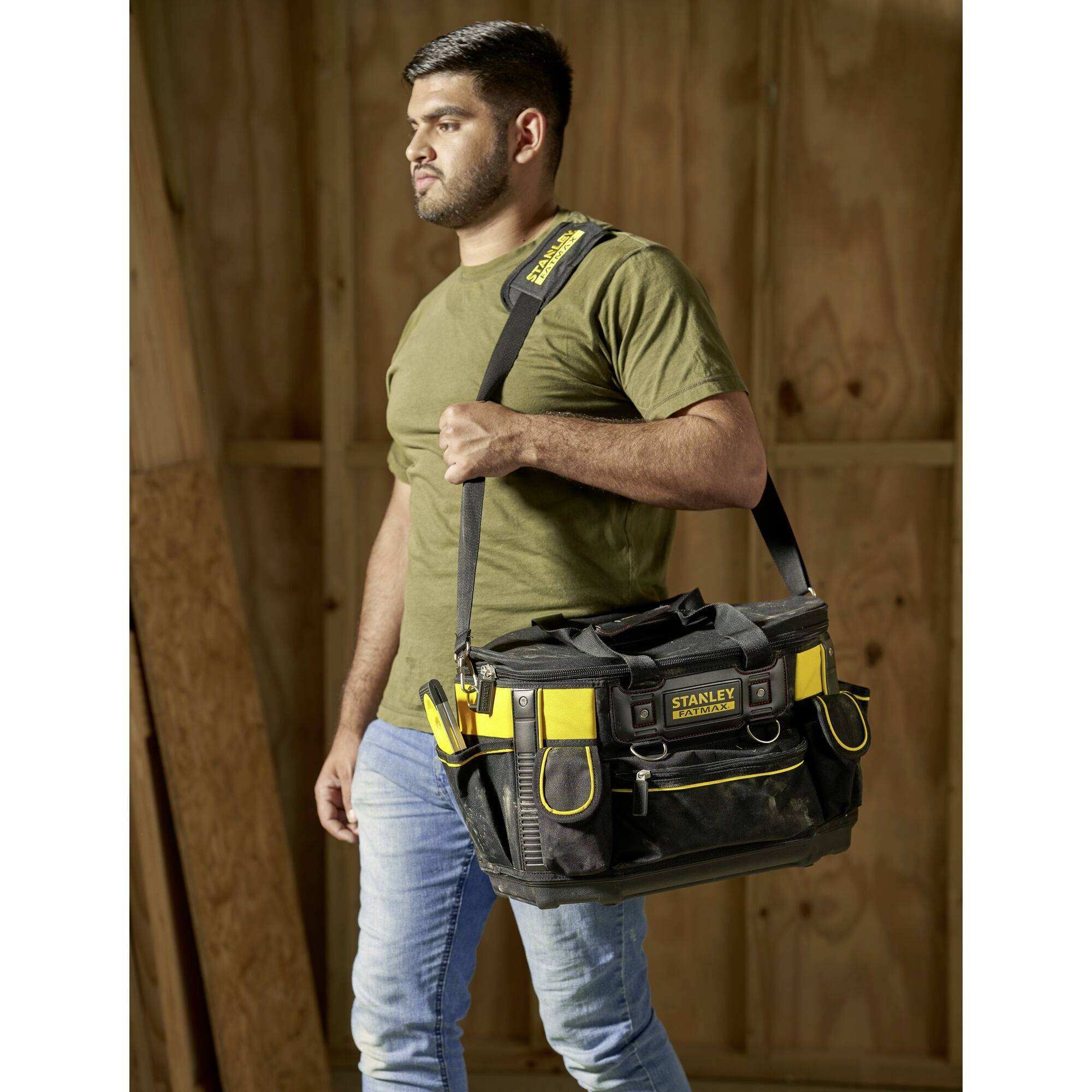 A man is carrying a black tool bag with yellow accents. The bag has multiple compartments and the man is wearing an olive green T-shirt.