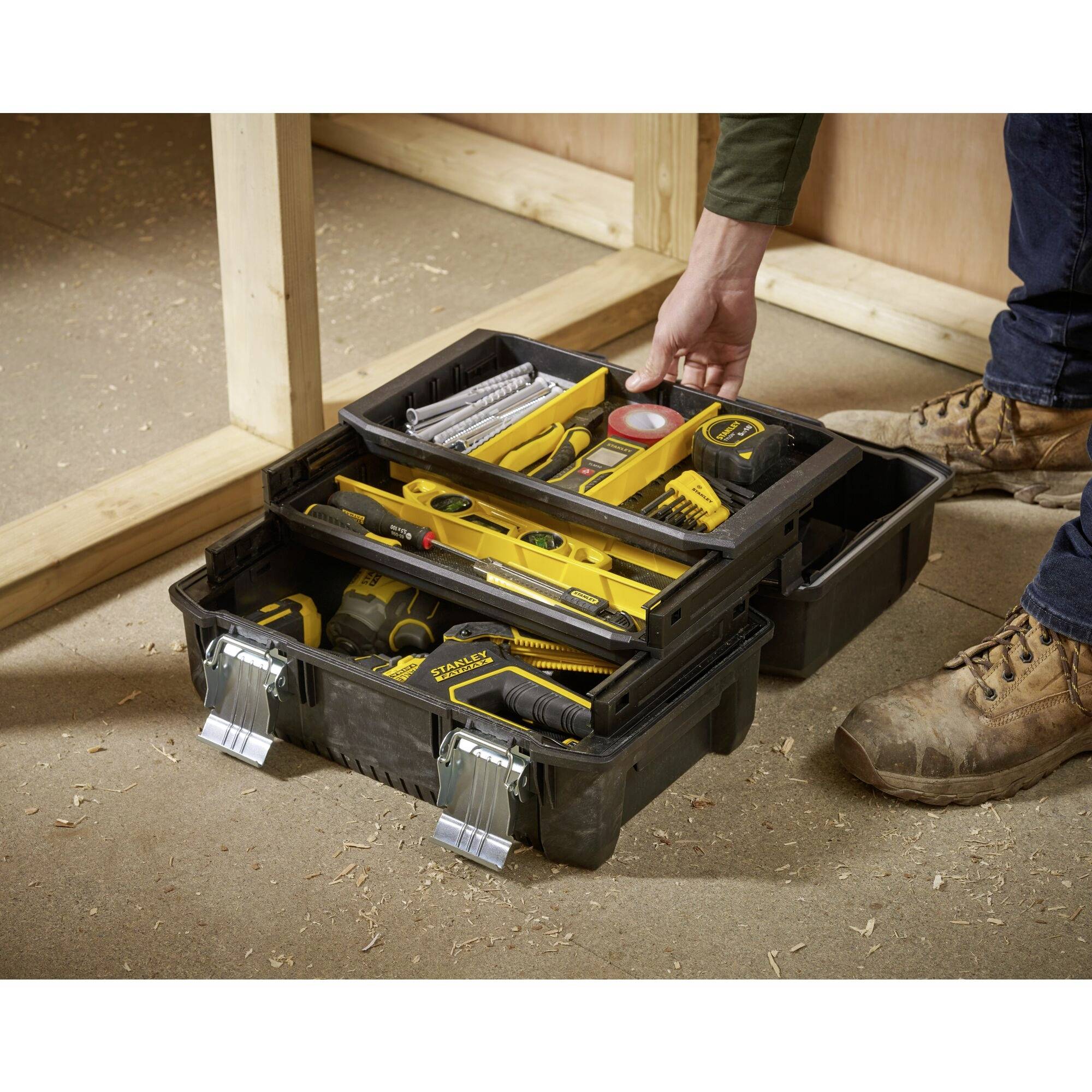 An open toolbox on the floor with yellow hand tools and accessories. A person in workwear is reaching for tools.