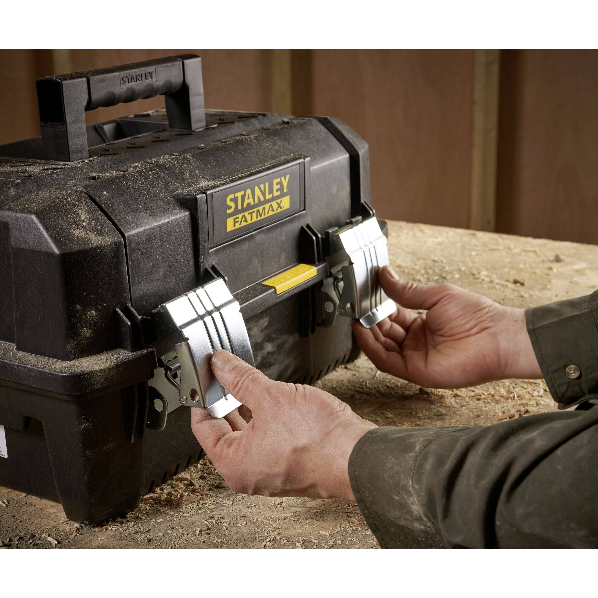 A person opens the latch of a 'Stanley Fatmax' toolbox on a wooden workbench.