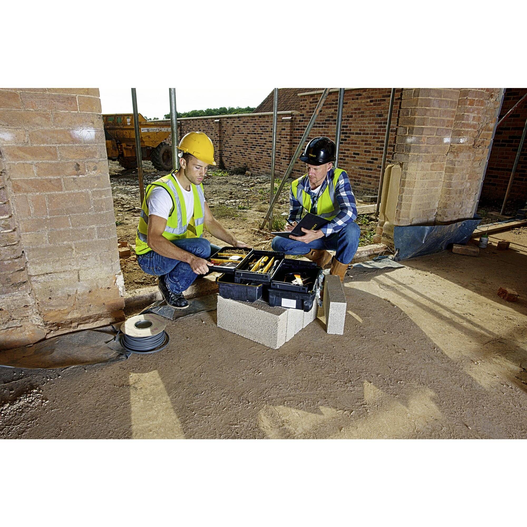 Two construction workers in hard hats and high-visibility vests are reviewing building plans on a building site next to a wall with construction materials in the background.