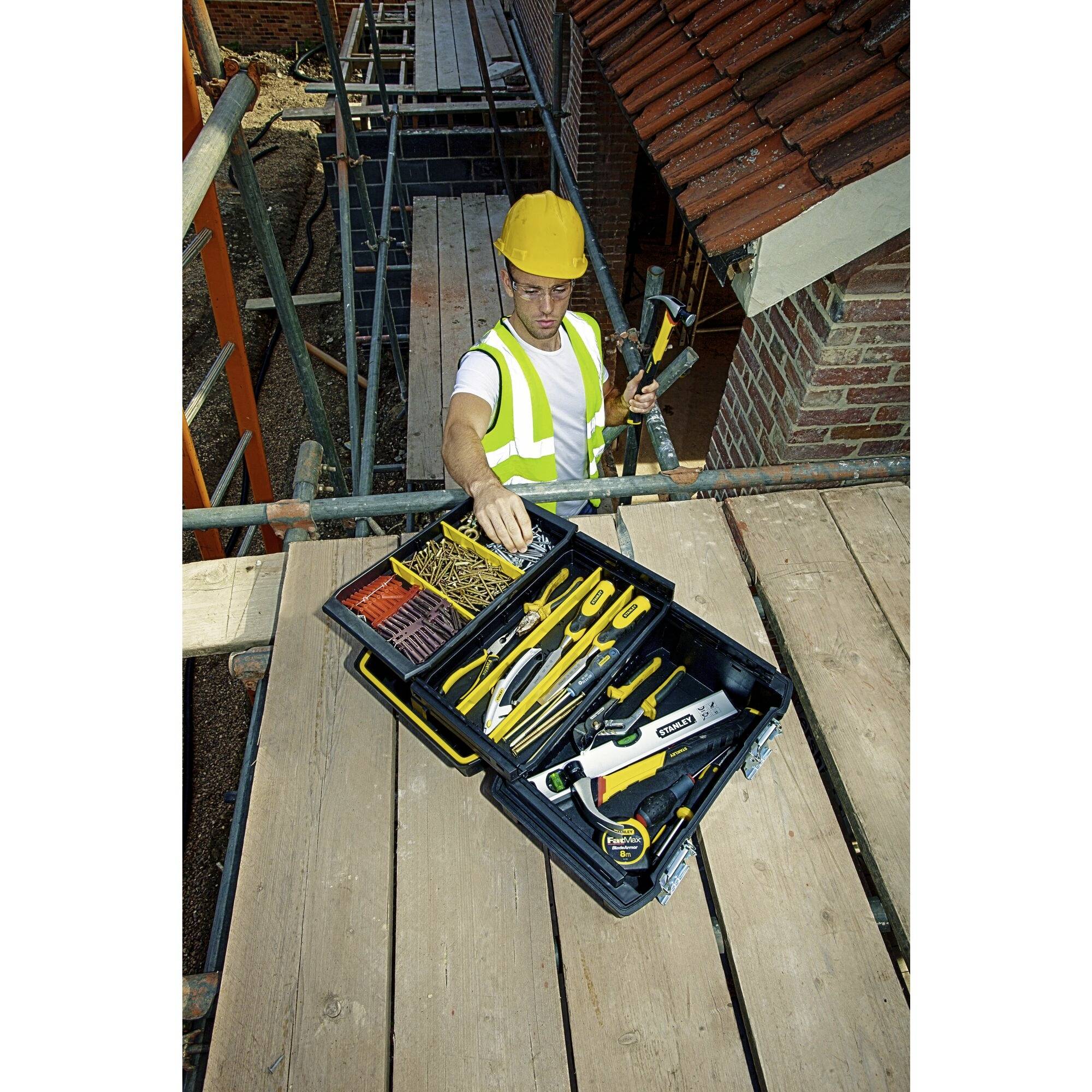 A construction worker in protective clothing stands on scaffolding and removes a tool from an open toolbox.