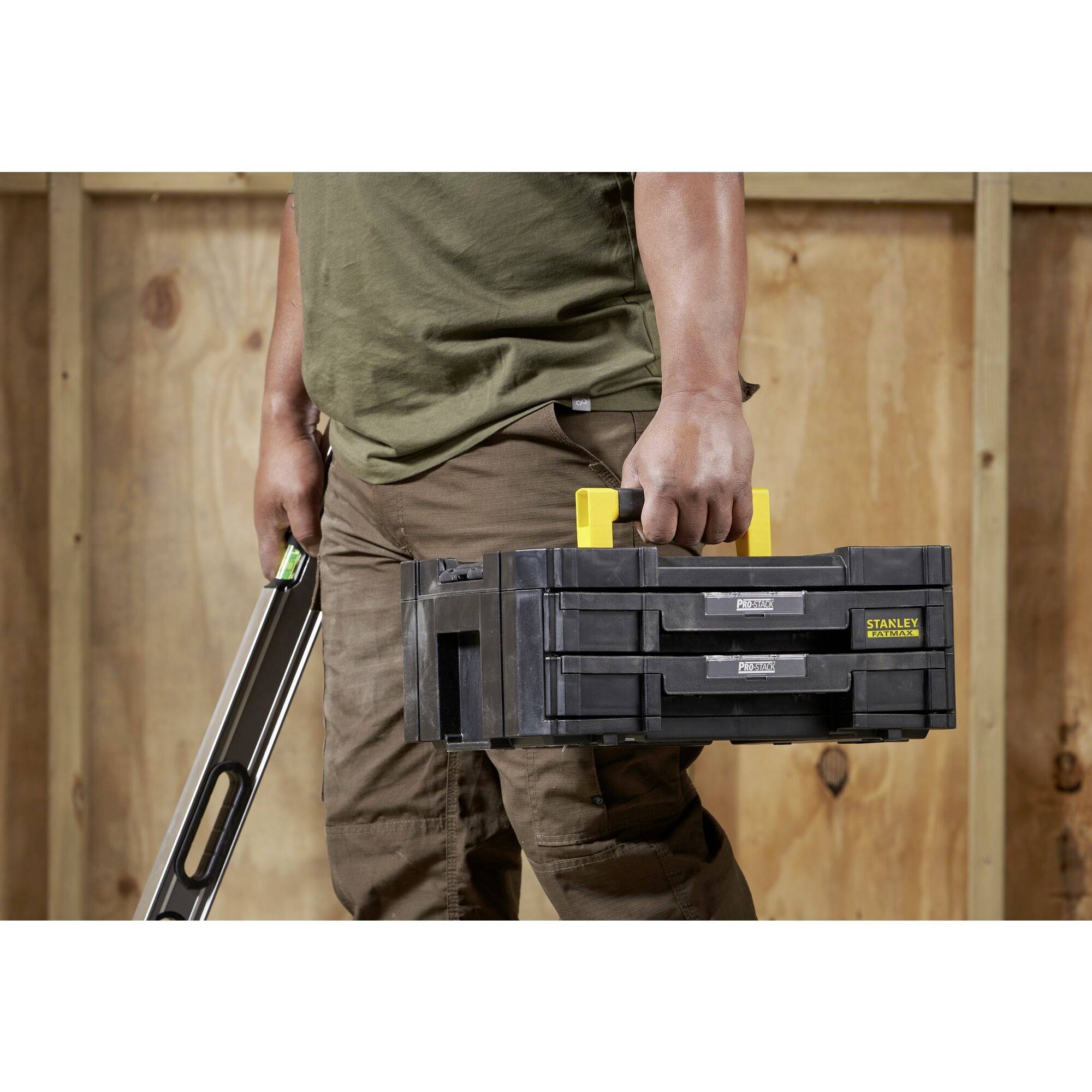 A person is carrying a black toolbox with a yellow handle and holding a spirit level. The background consists of wooden boards.