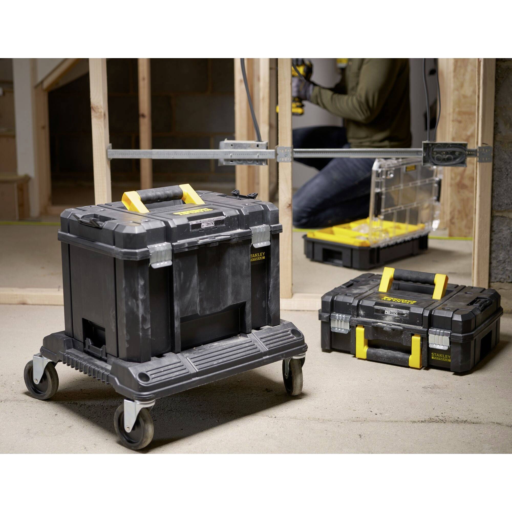 Two black tool cases with yellow handles are standing on a workshop floor. A craftsman in the background is working on a project.