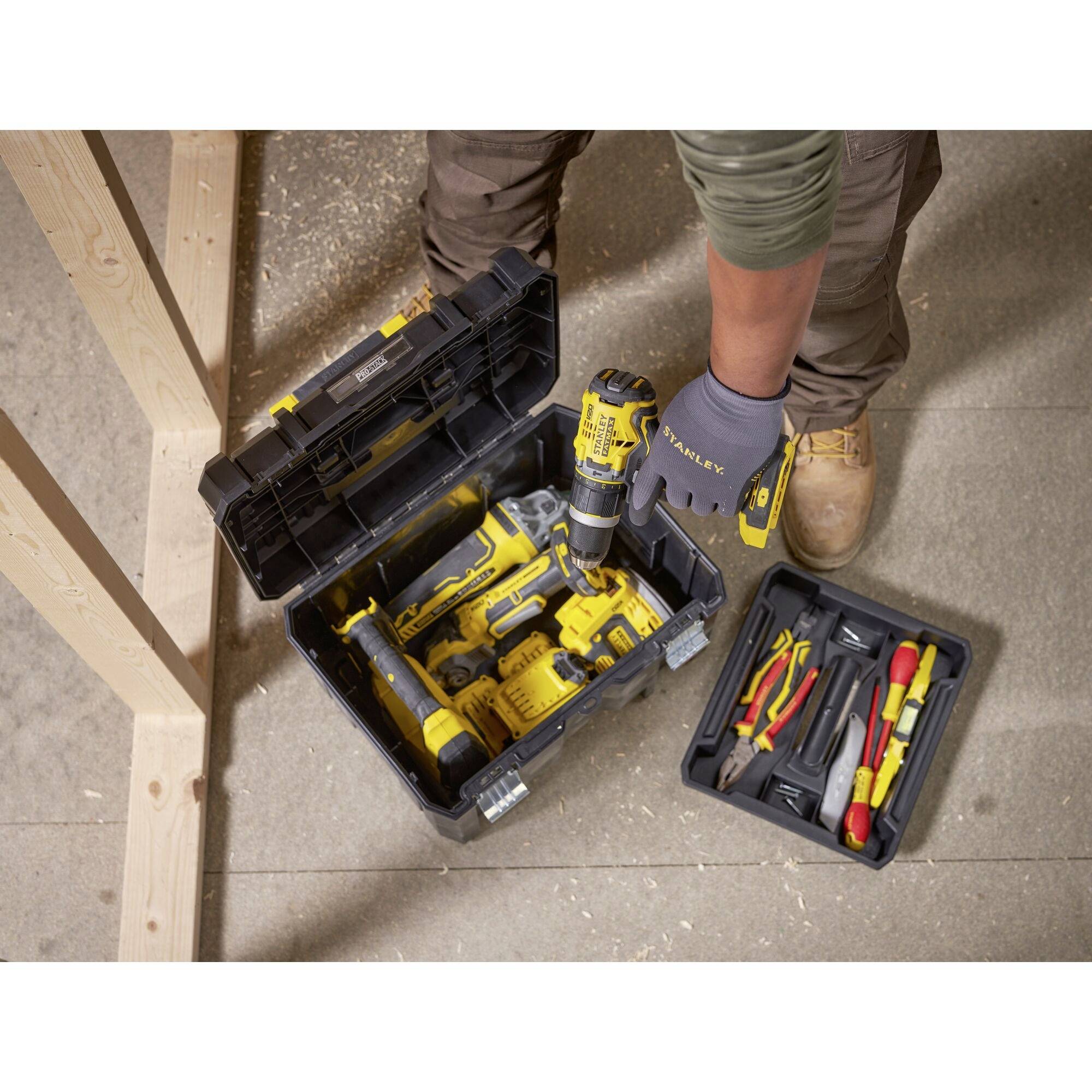 A person is standing on a wooden floor, holding a cordless drill above an open toolbox containing various tools.