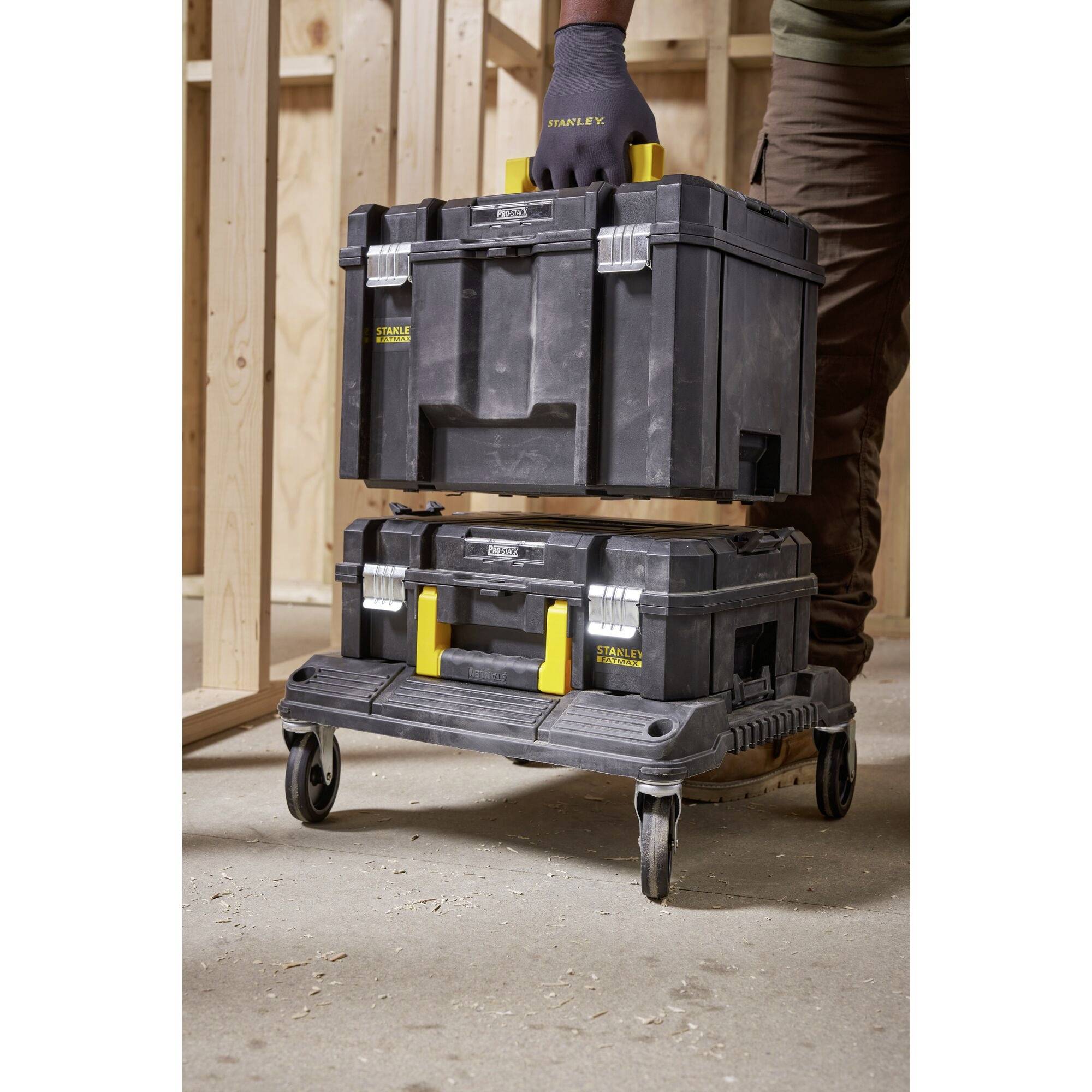 A person is stacking two black tool cases with yellow handles, with the top one being lifted, wooden environmental frame in the background.