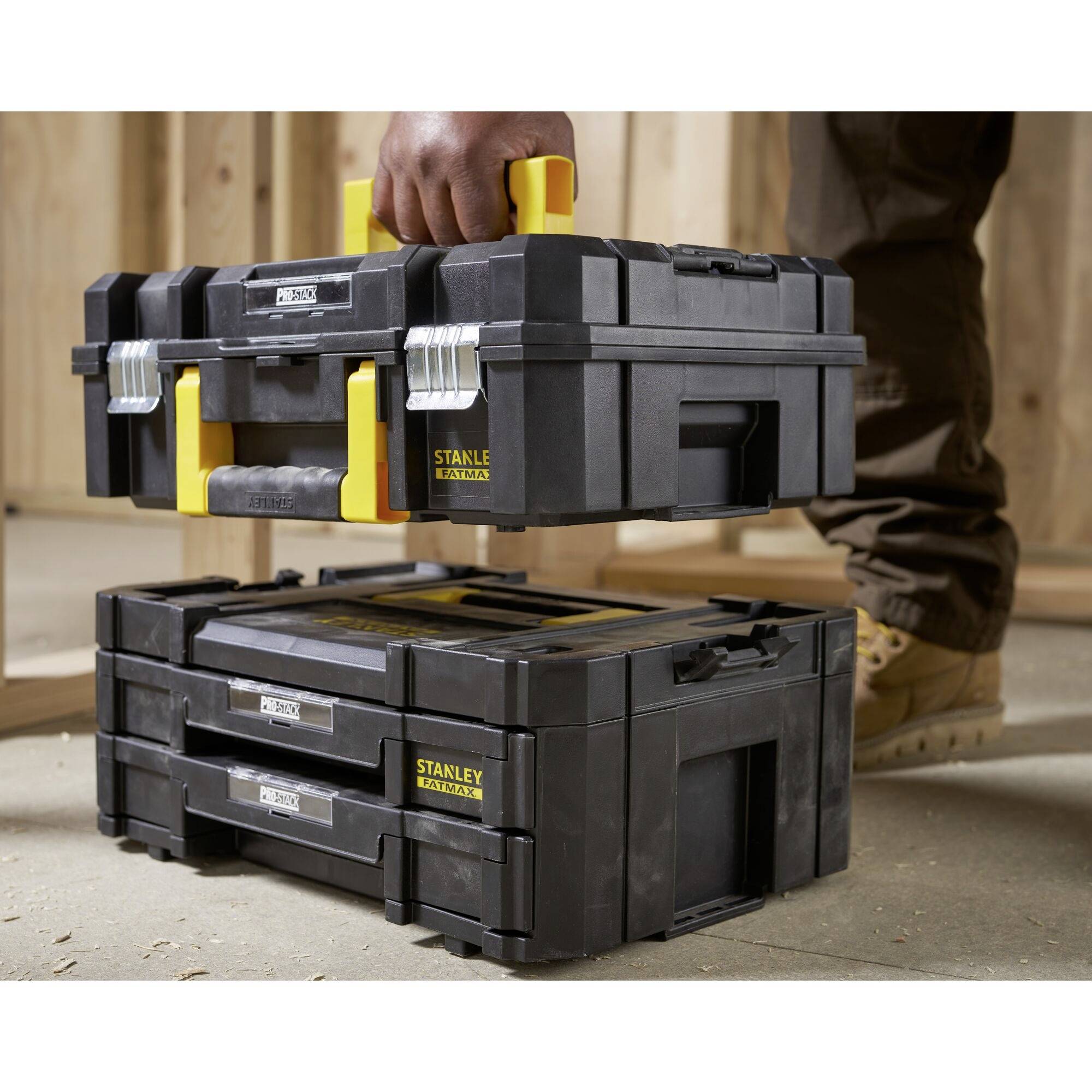 A tradesman is lifting a black, stackable toolbox with a yellow handle. Two similar boxes are standing on a concrete floor.