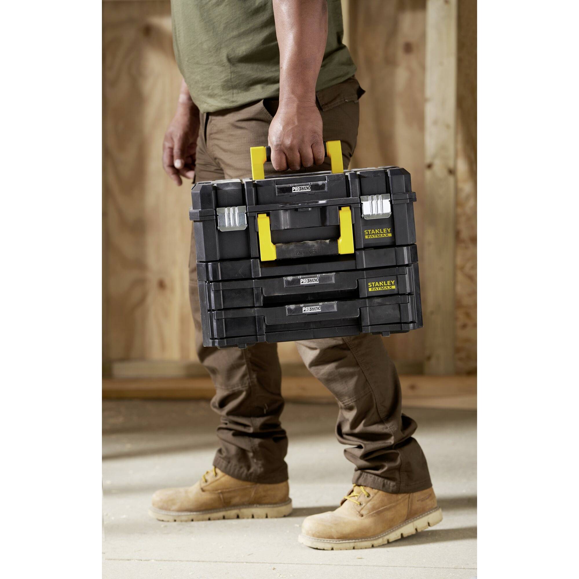 A person is carrying a black toolbox with a yellow handle in a workshop environment. The walls are unfinished.