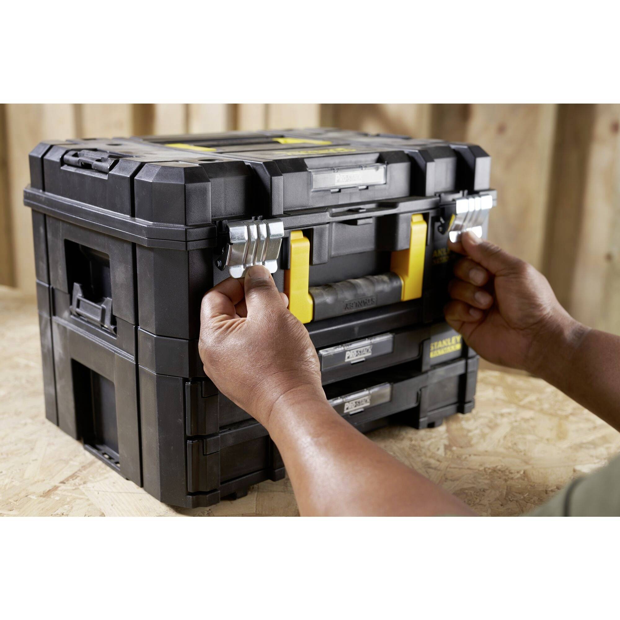 A person opens a black toolbox on a wooden surface, using both hands to lift the side latches.
