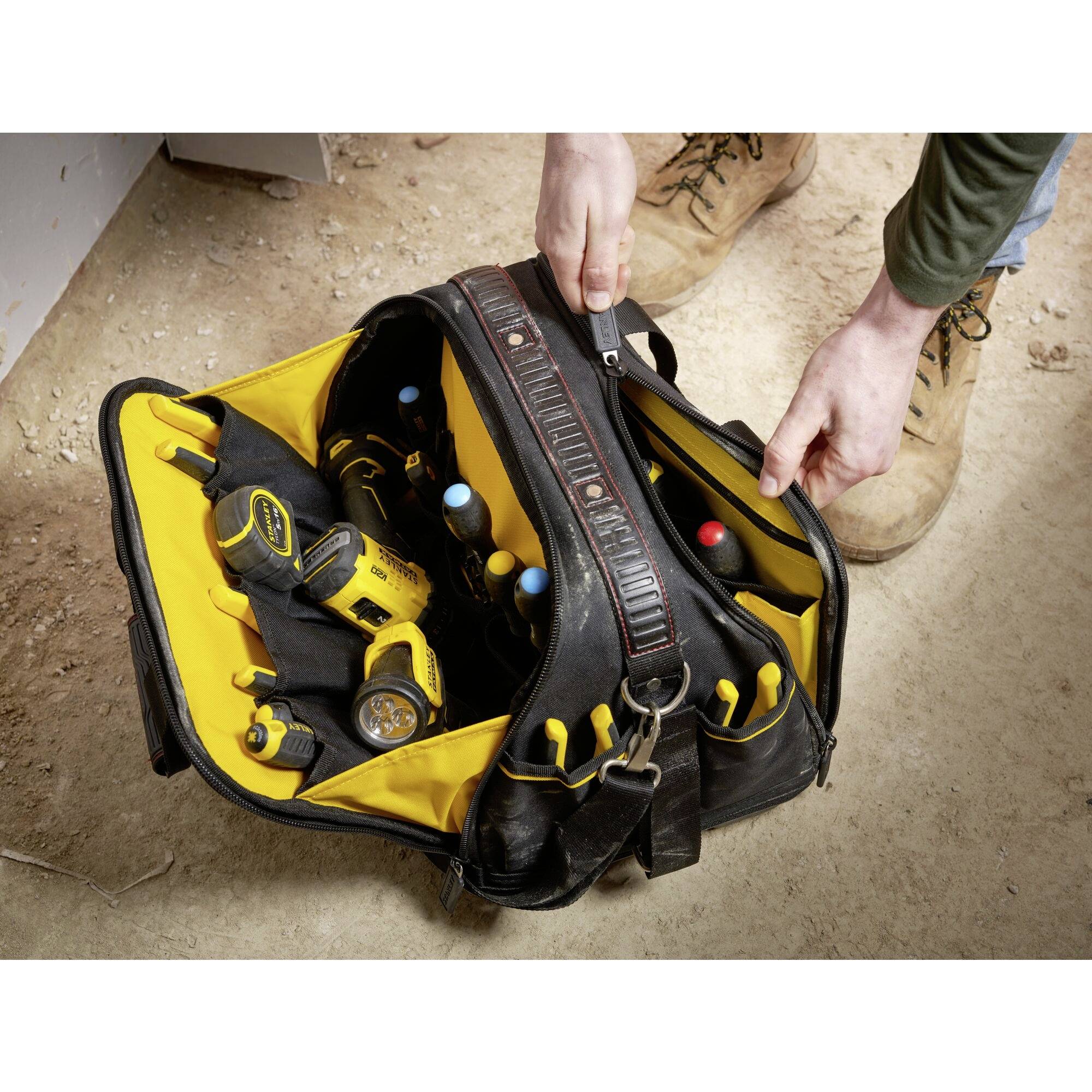 An open tool bag on a floor displays electrical tools and screwdrivers. A person reaches for a tool.