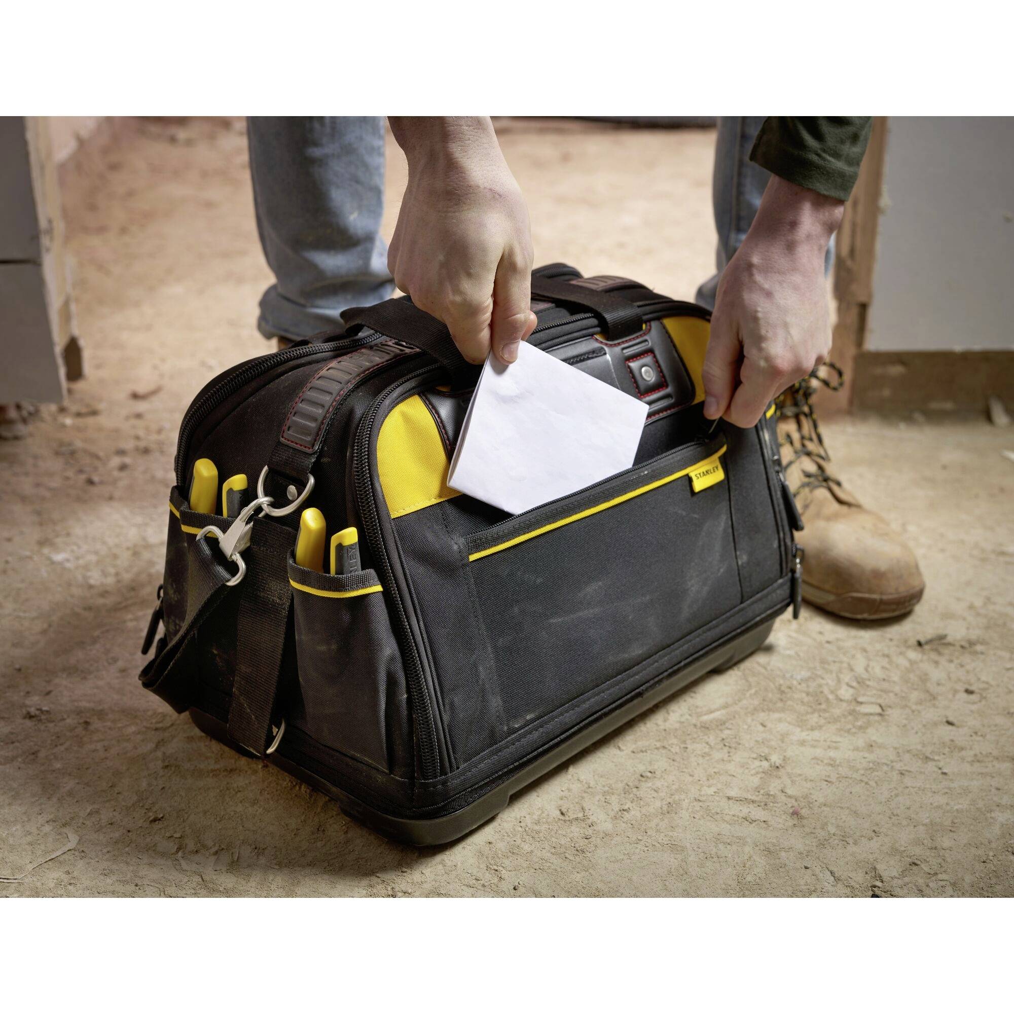A person is holding a black tool bag with yellow accents and is placing a white envelope inside. The background shows a construction site environment.