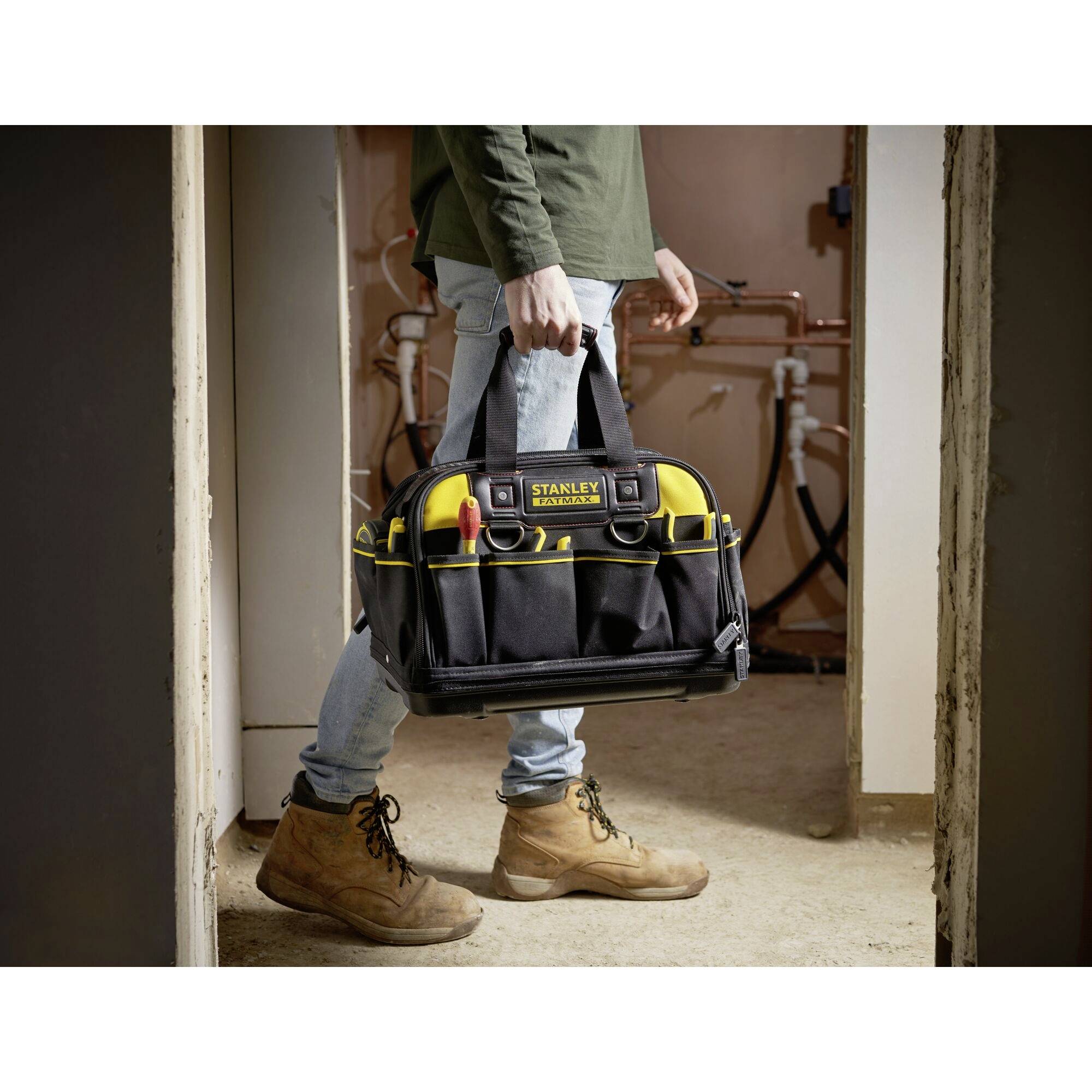 A worker in jeans, boots and a green shirt is carrying a filled tool bag through a room with unfinished walls and exposed pipework.