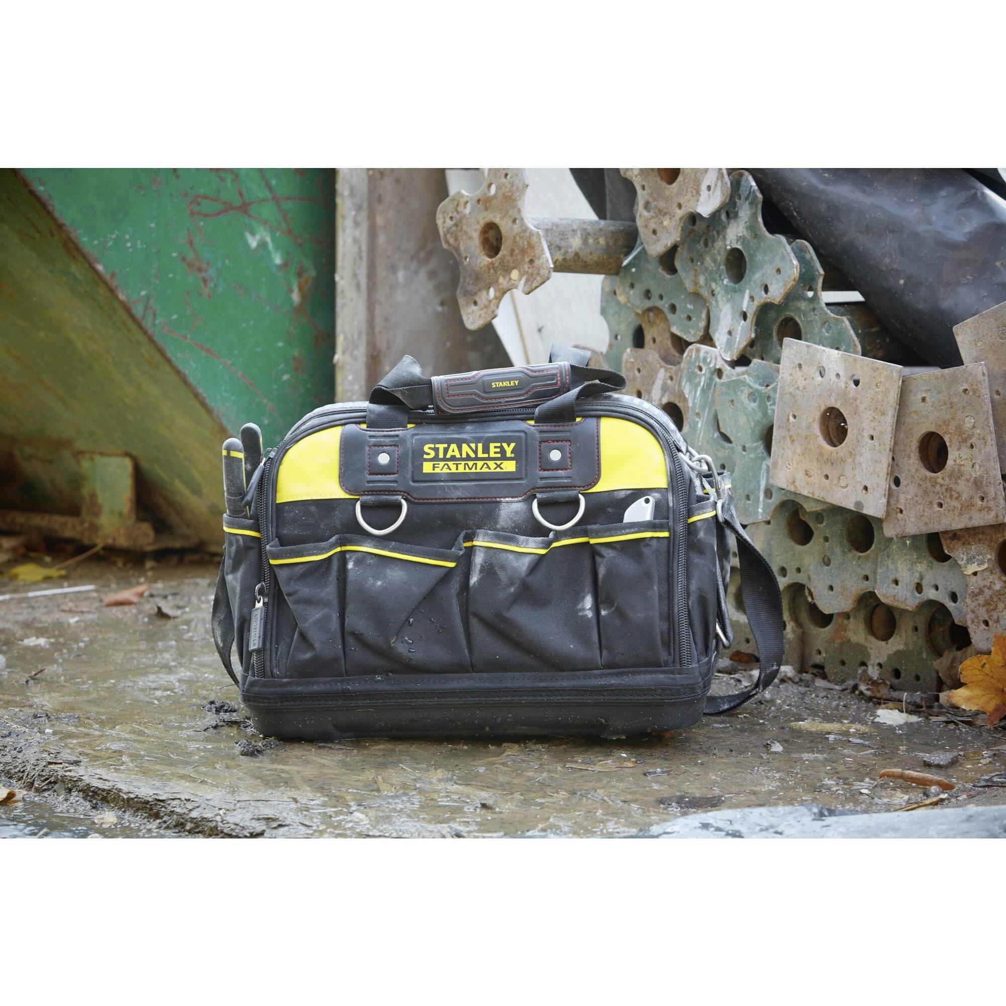 'Black tool bag with yellow accents and multiple pockets, standing on wet ground in front of a green container and concrete blocks.'